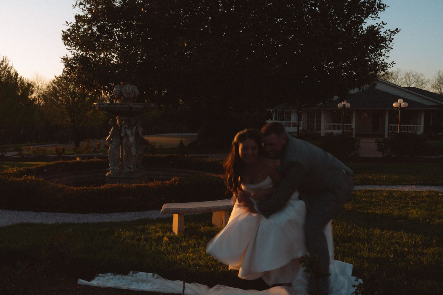 Couple sharing a romantic dip kiss at golden hour in a garden with a fountain and bench at a wedding venue.