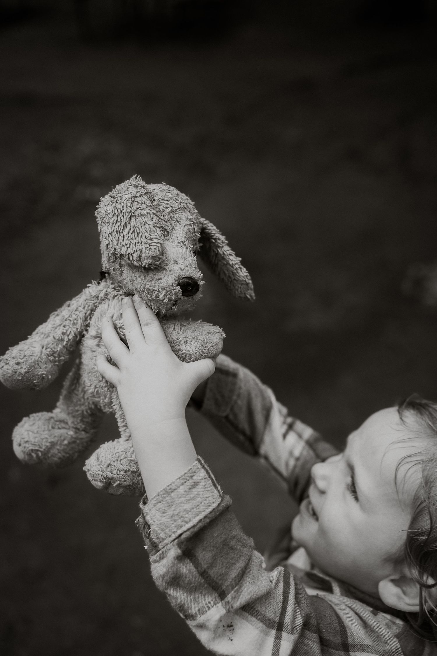 Black and white photo of a young child holding up a stuffed dog toy outdoors.