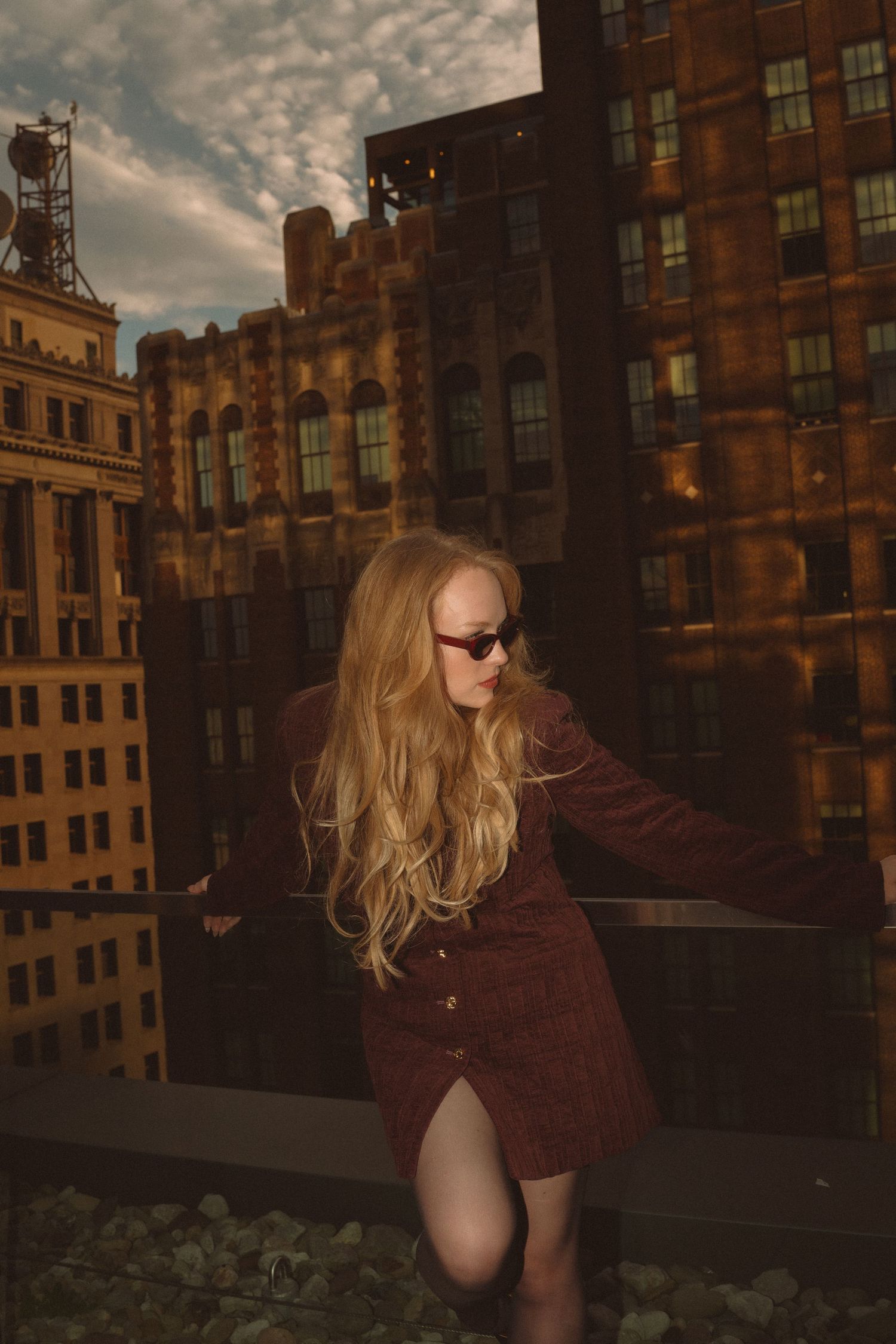 Blonde woman in burgundy dress and sunglasses poses on urban rooftop with brick buildings backdrop.