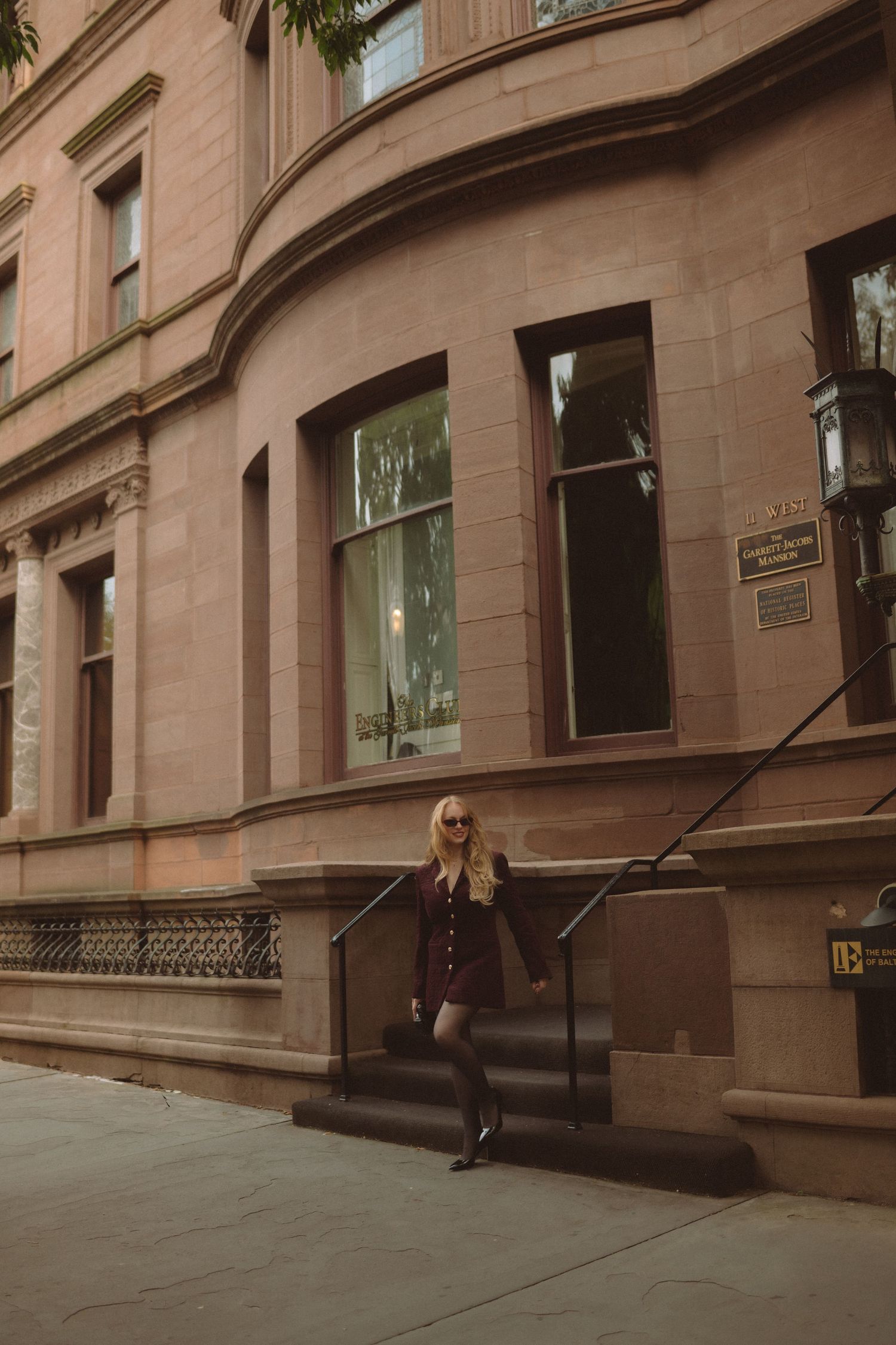 Blonde woman in dark outfit poses on steps of a classic brownstone building on a city street.