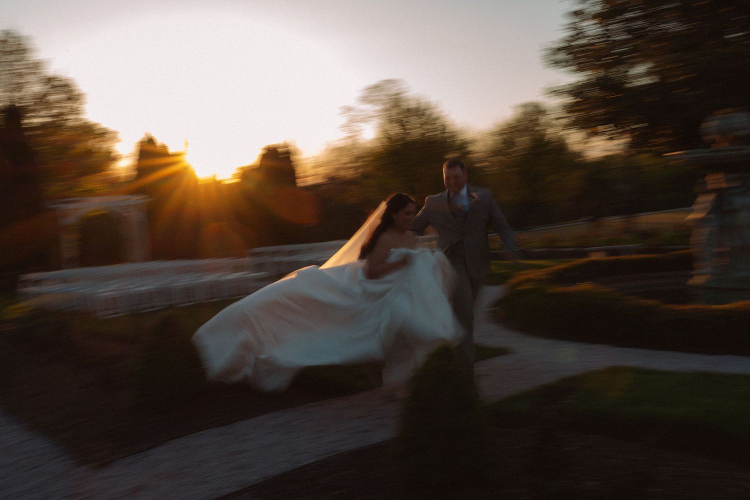 Bride and groom running at sunset in a garden, motion blur capturing romantic golden hour wedding moment.