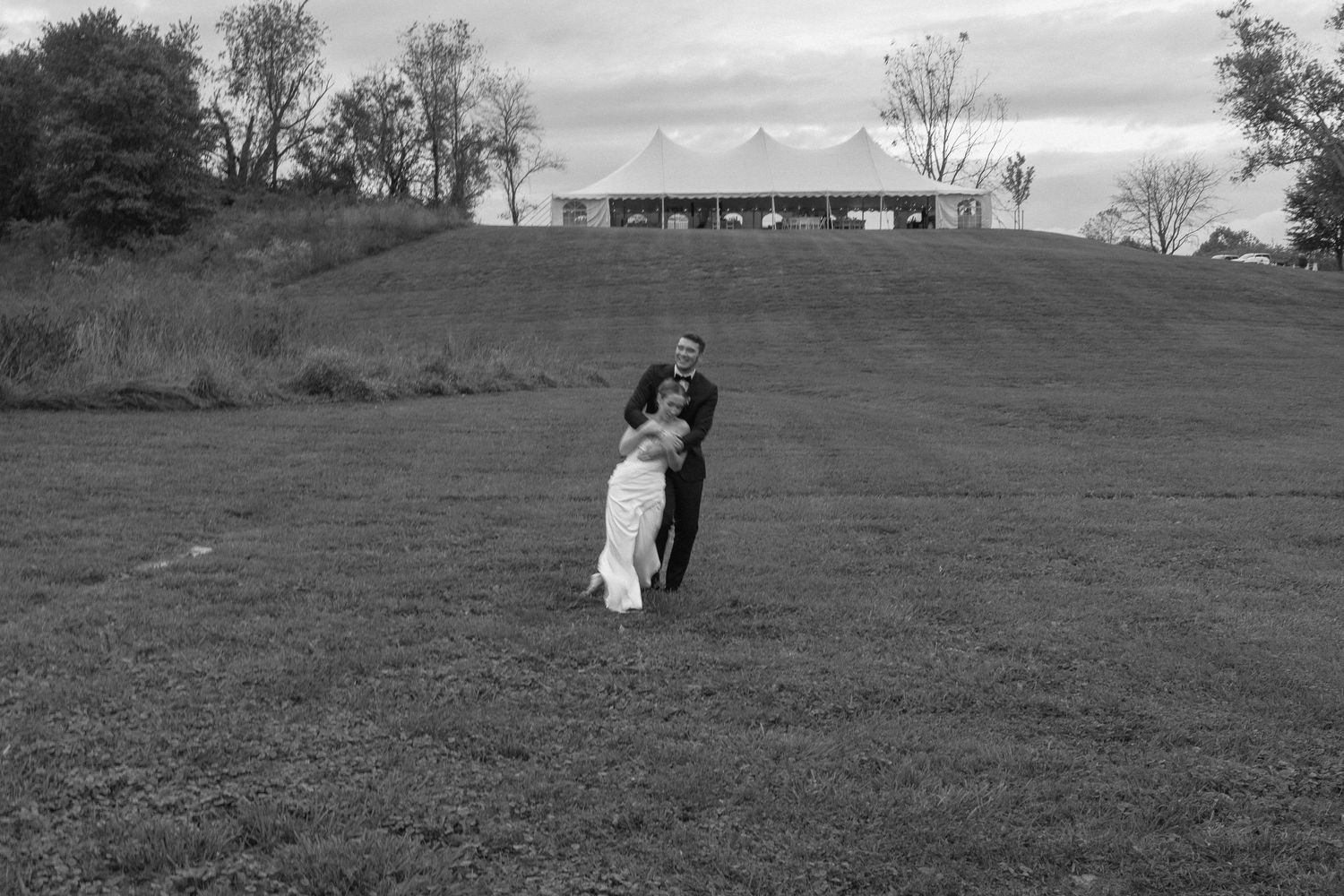 Black and white photo of a couple embracing on a grassy hill with a wedding tent in the background.