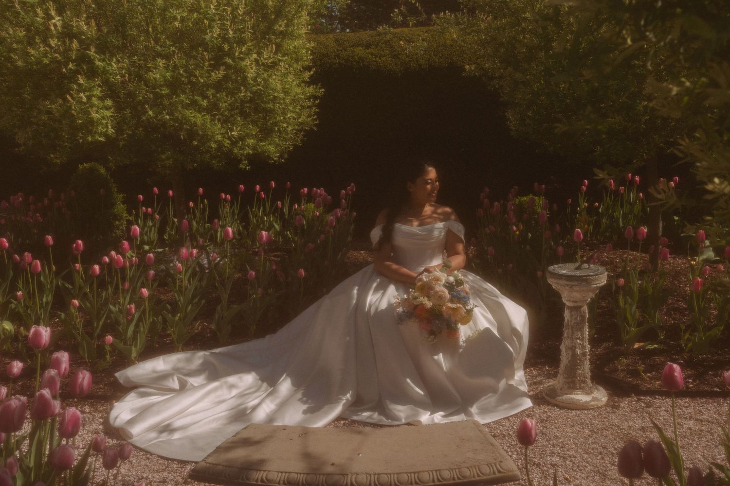Bride in white ball gown sitting in a sunlit garden surrounded by pink tulips, holding a bouquet.