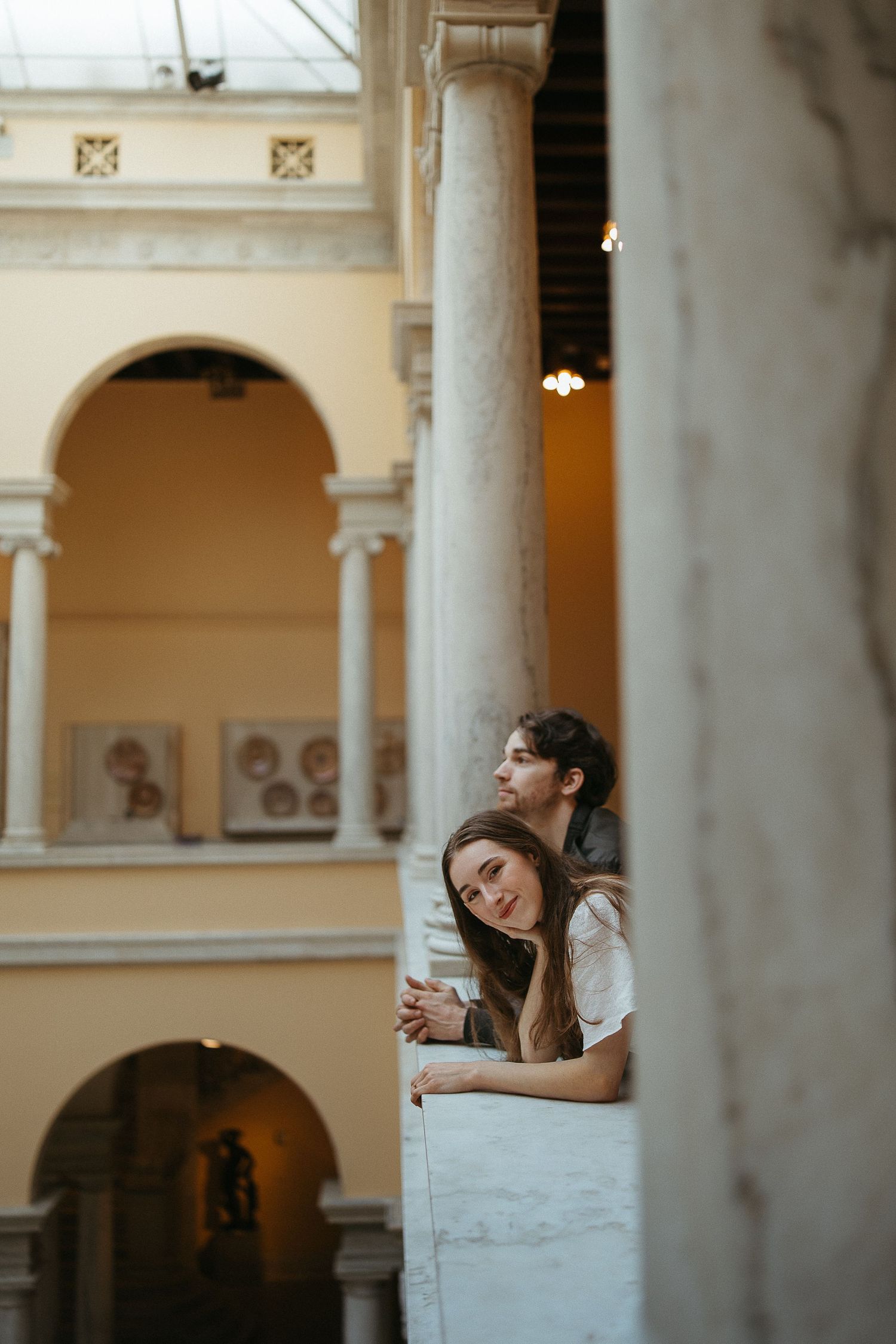 Two women leaning on a marble balcony railing inside a classical museum with arched ceilings and columns.