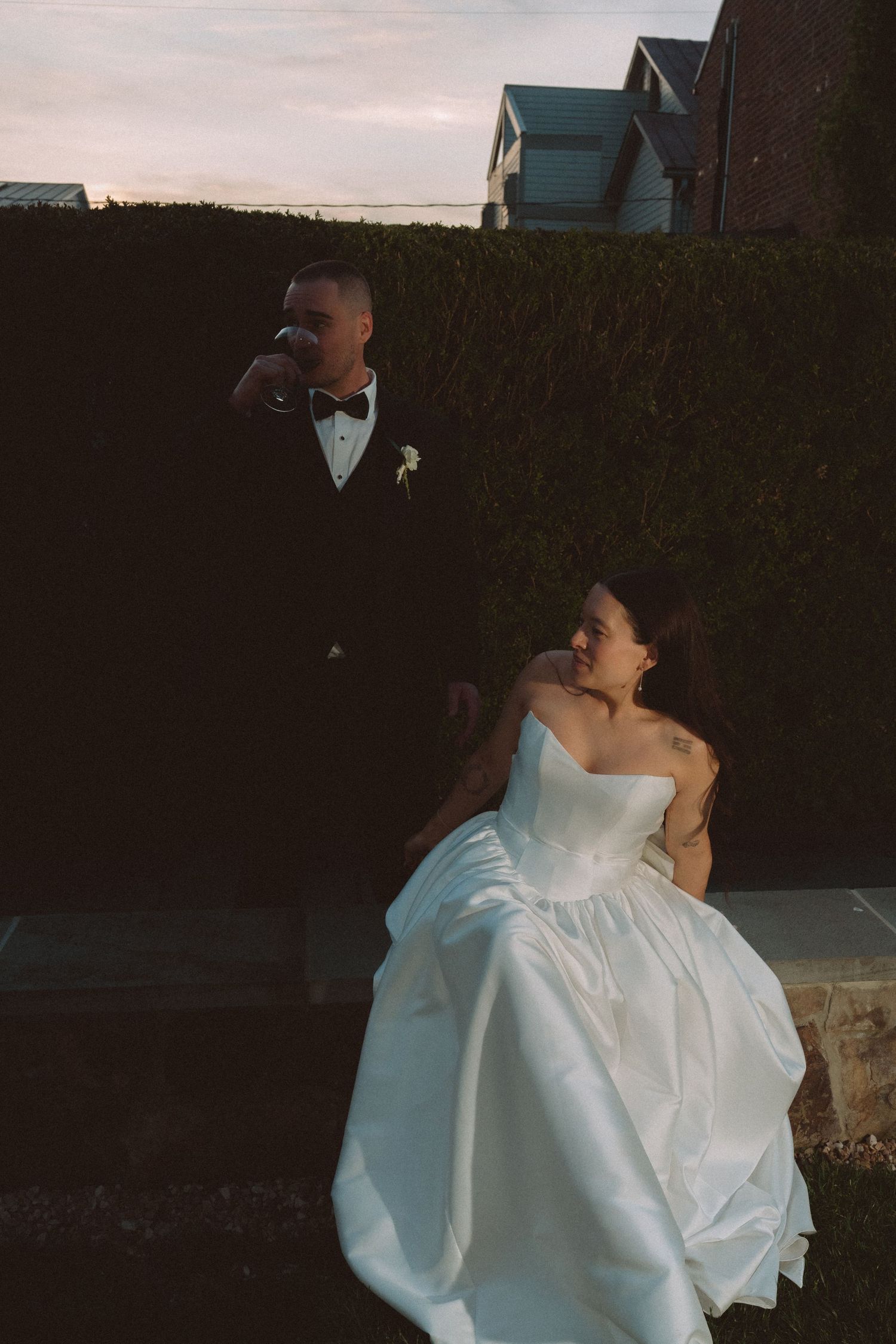 Bride in strapless white ball gown sits beside groom in black tuxedo drinking wine at twilight near hedge.