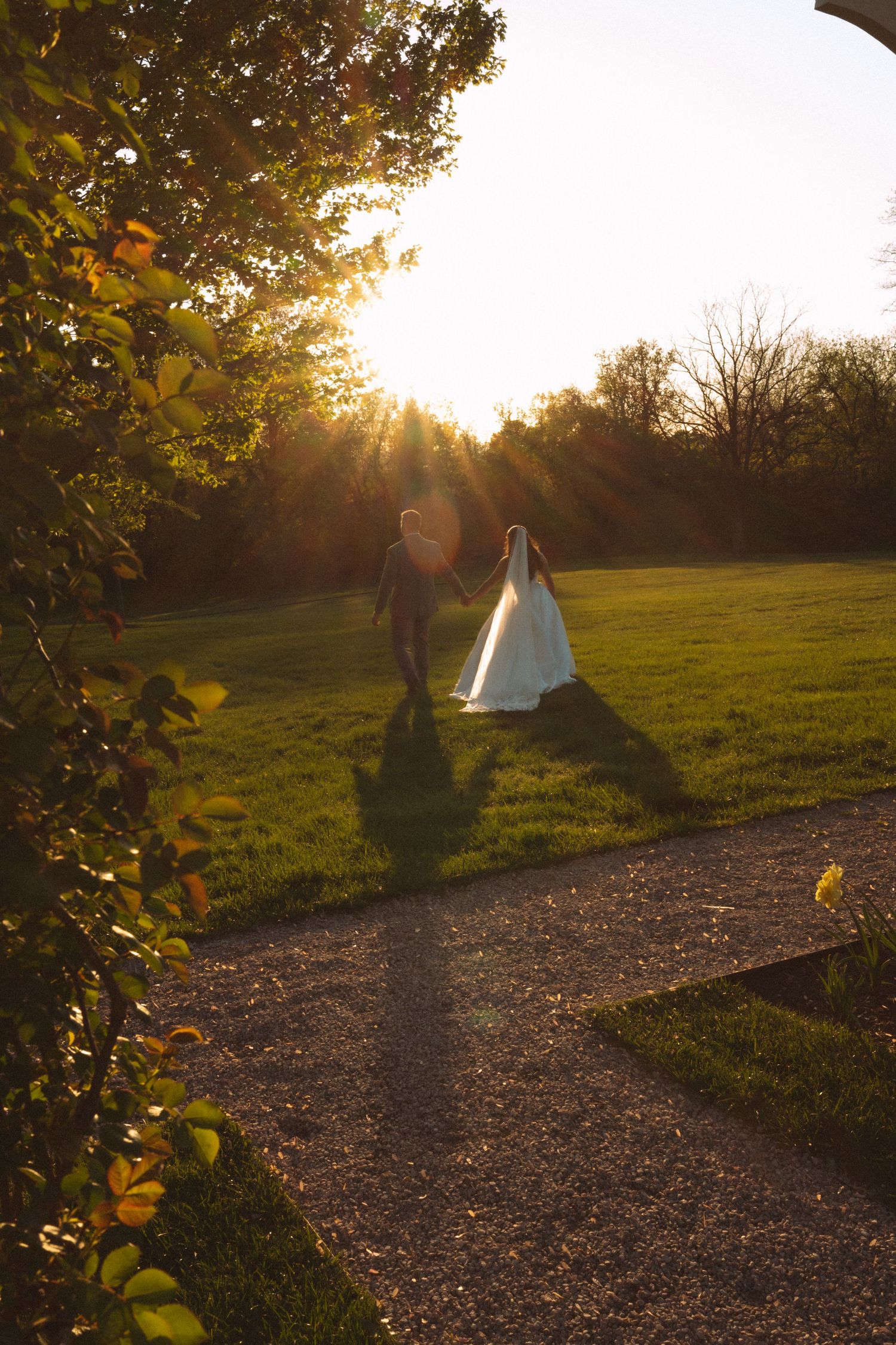 Bride and groom walking hand in hand on a garden path during golden sunset at an outdoor wedding.