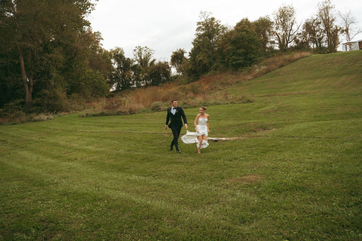 A bride and groom walk hand in hand across a lush green hillside at their outdoor wedding ceremony.