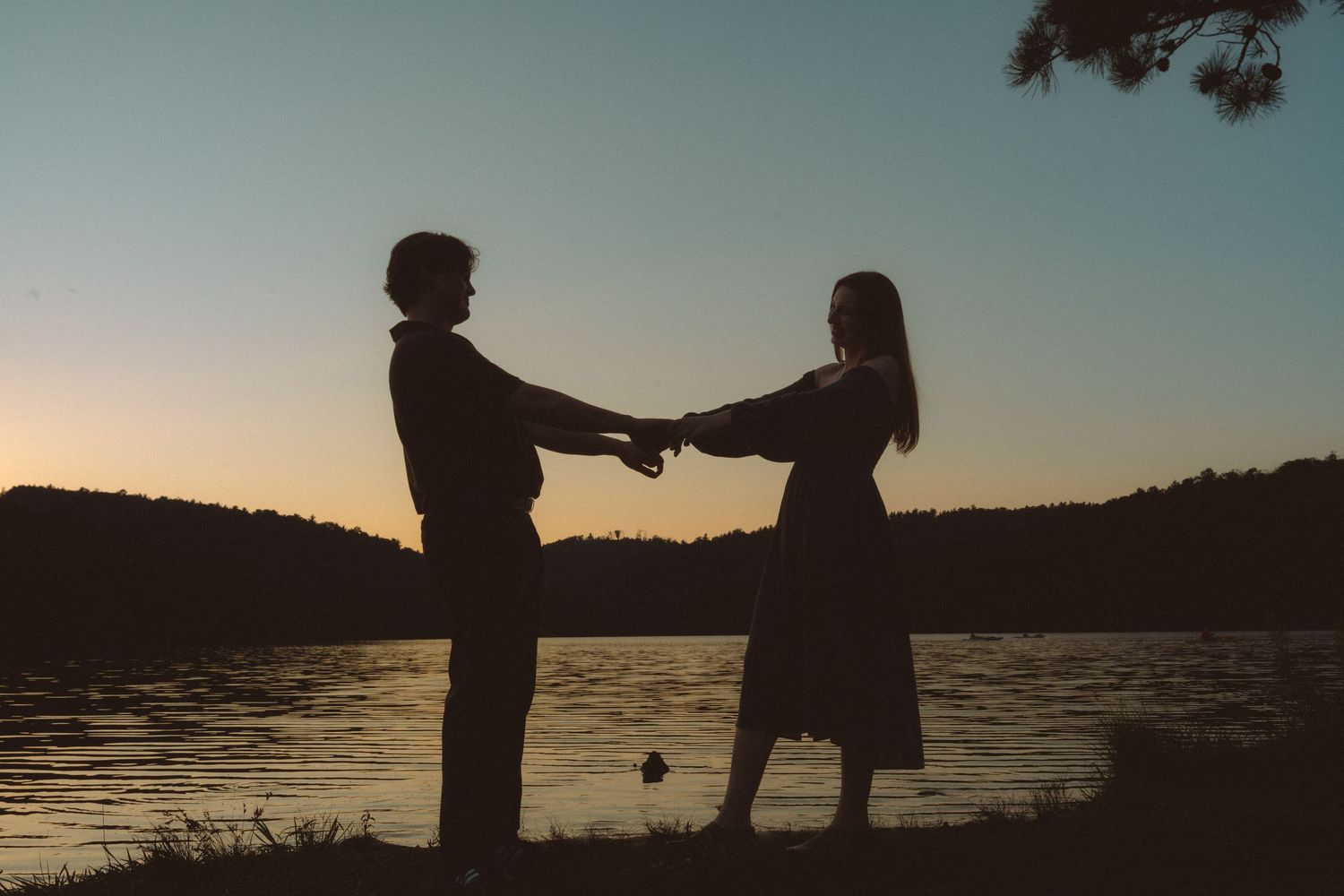 Silhouette of a couple holding hands by a lake at sunset with trees in the background.