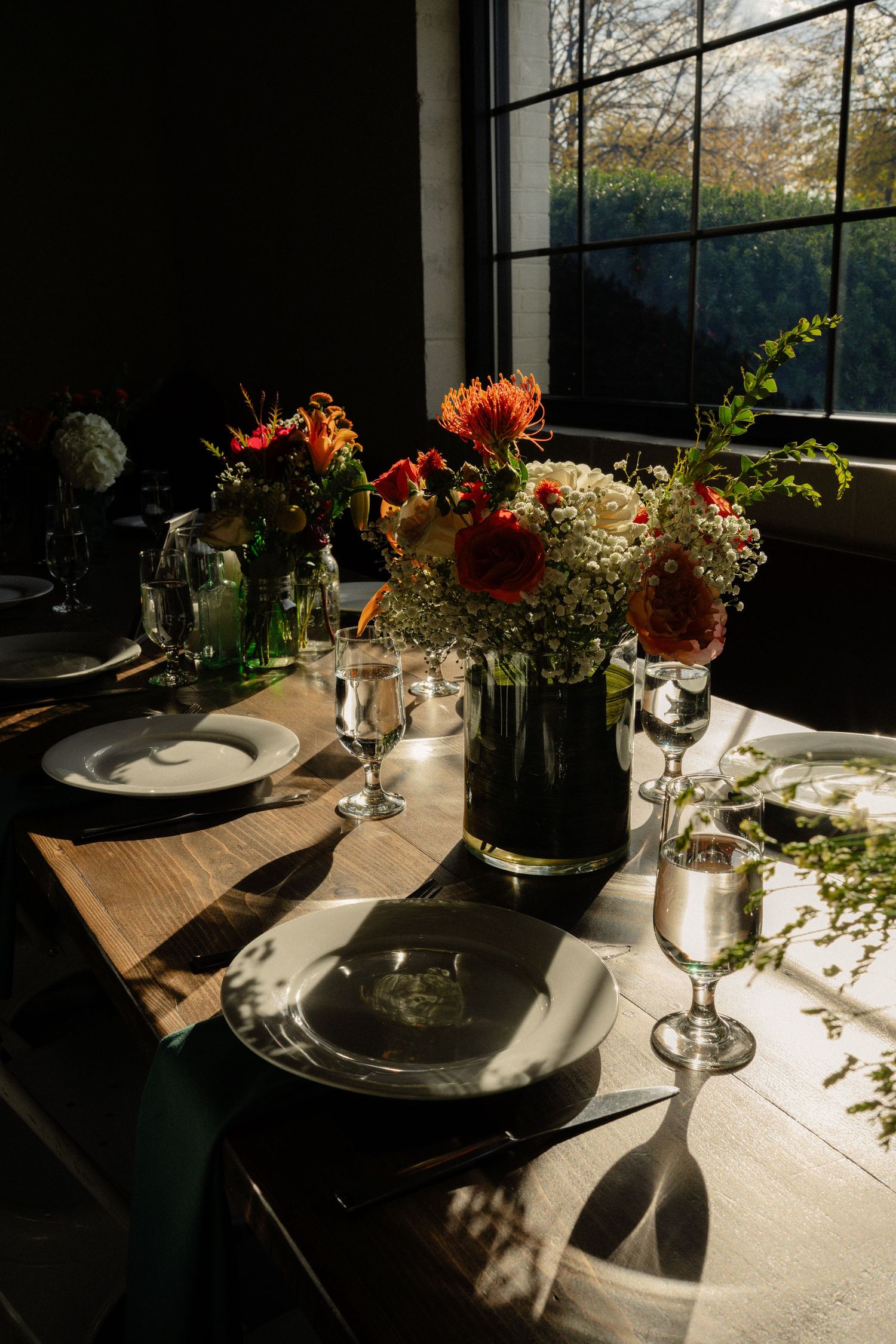 Elegant dining table set with white plates, crystal glasses, and orange floral centerpiece bathed in natural window light.