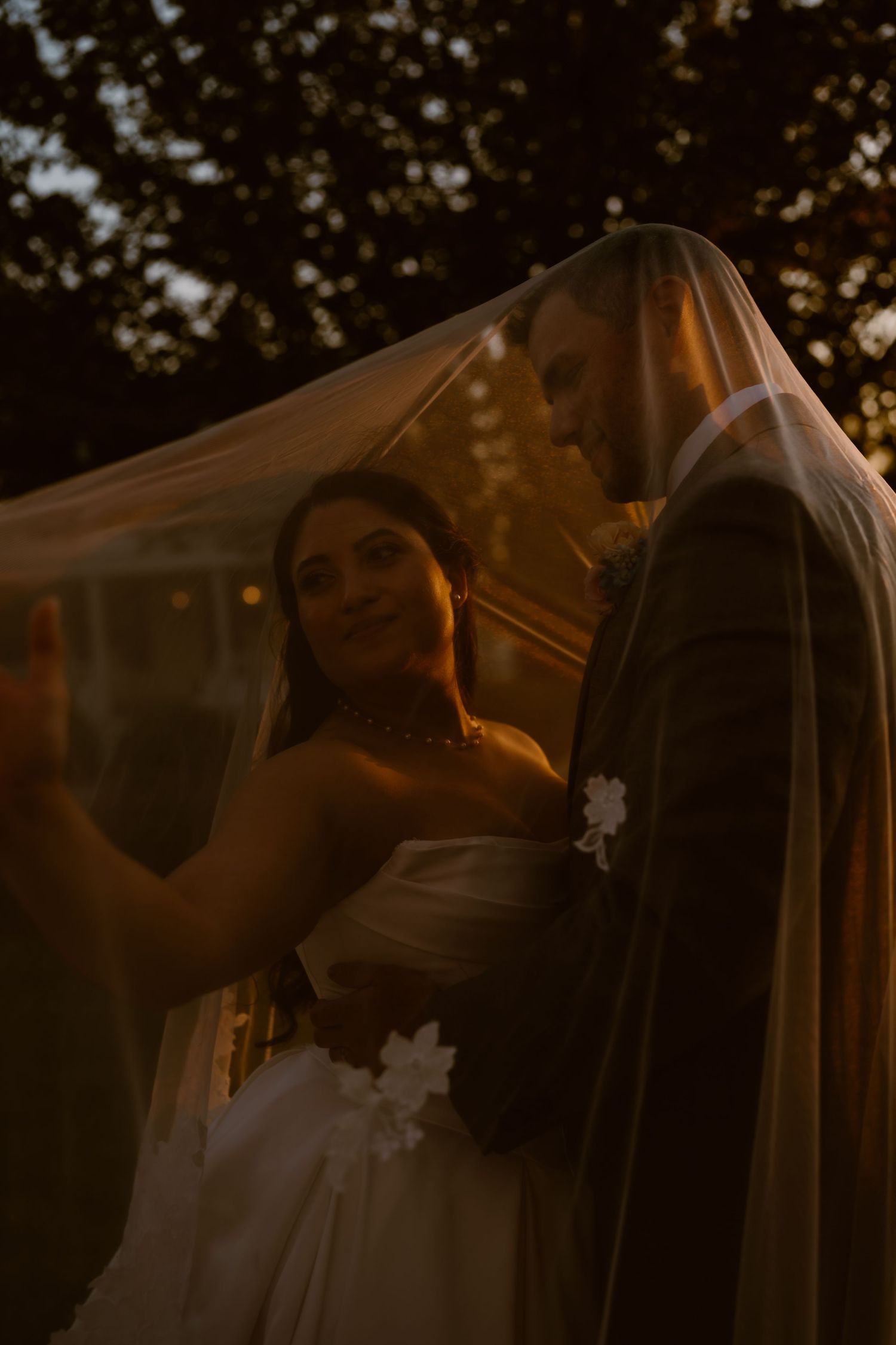 Bride and groom share romantic moment under sheer wedding veil at golden hour outdoor ceremony.