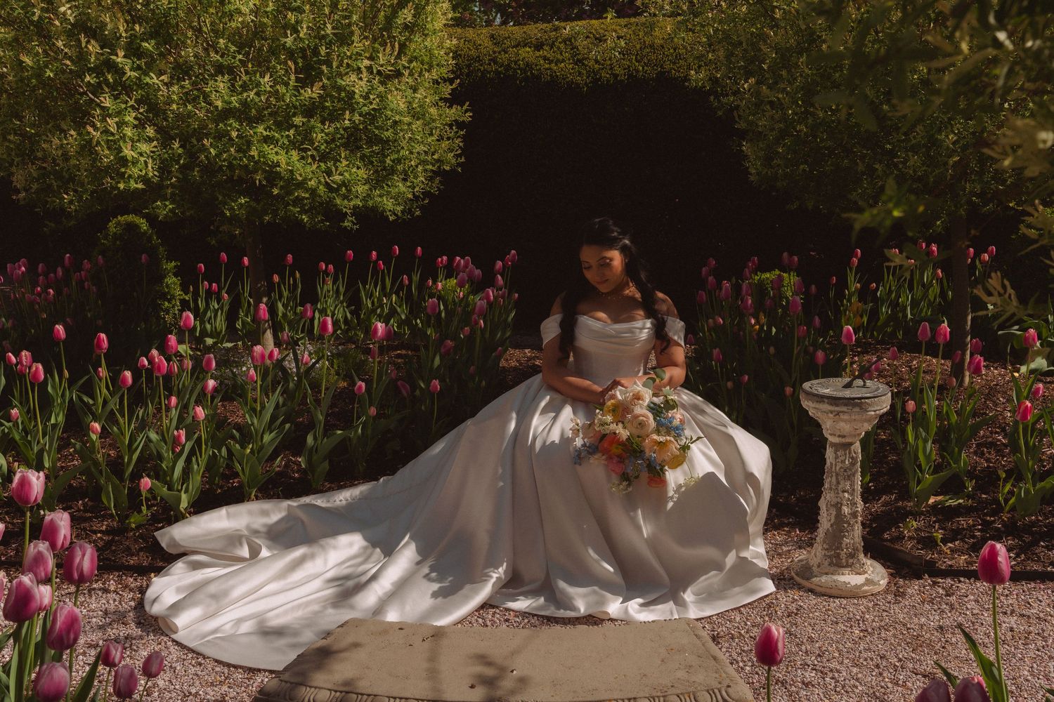 Bride in white gown sitting among pink tulips in a sunlit garden, holding a bouquet.