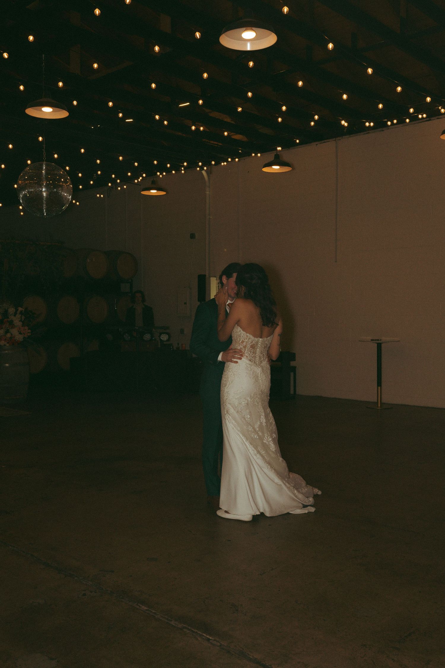 Bride and groom share first dance under warm string lights in a rustic winery wedding venue.