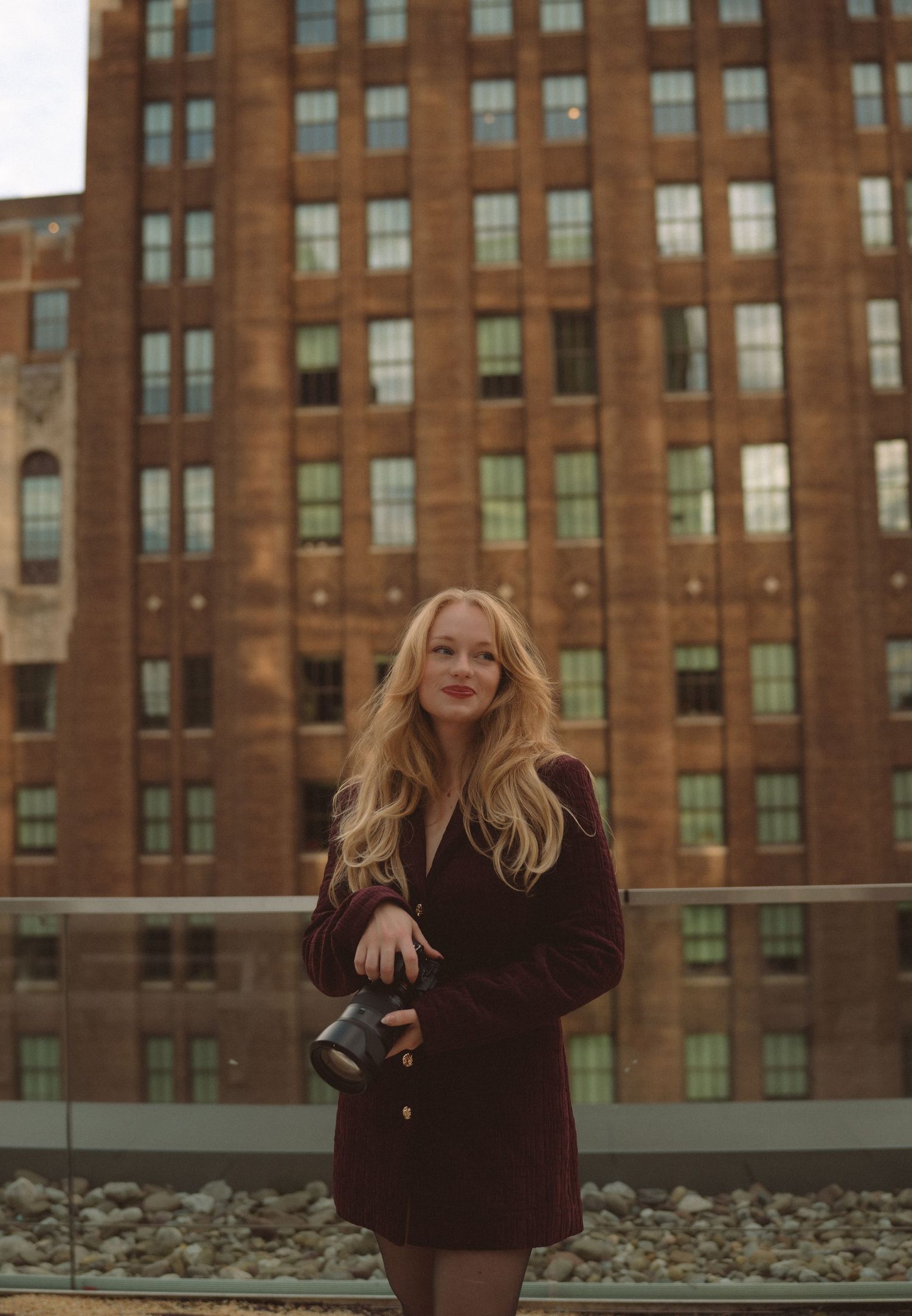 Blonde woman in burgundy coat holding a black bag, posing on a rooftop with a brick building backdrop.