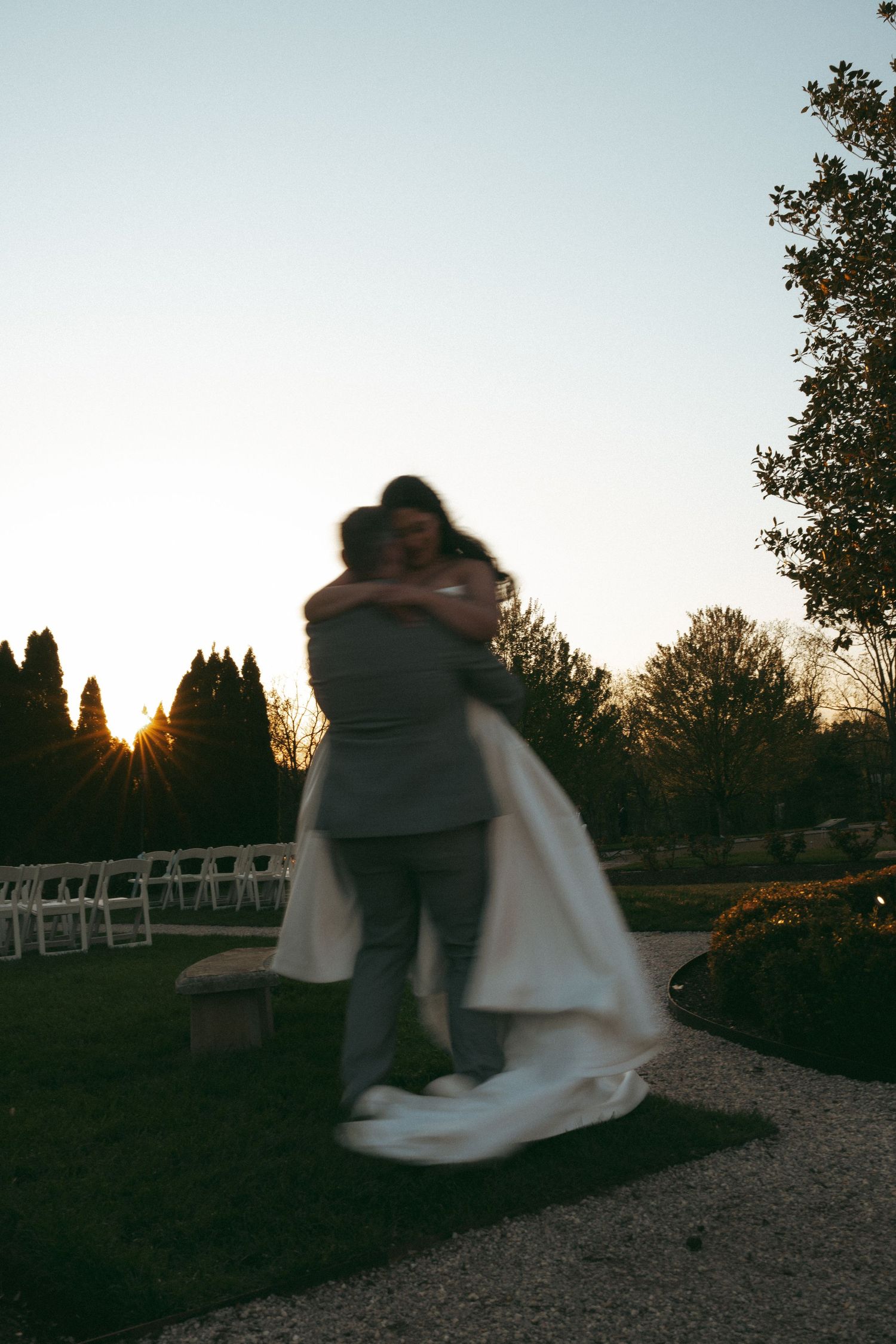 A couple embraces at sunset in a garden wedding venue, the bride in a flowing white gown being lifted by the groom.