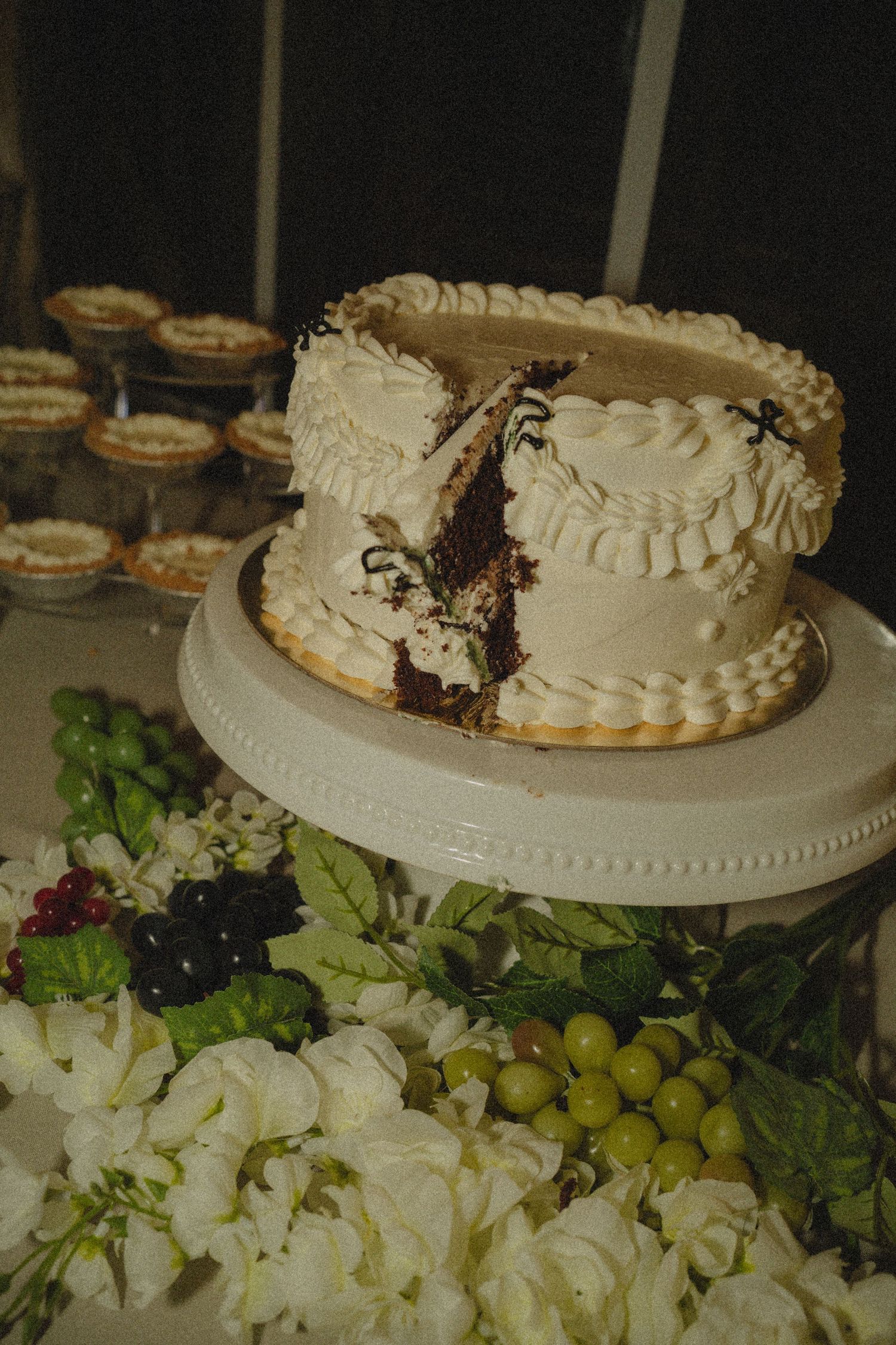 Sliced white frosted chocolate wedding cake on a stand, surrounded by white flowers and green grapes.