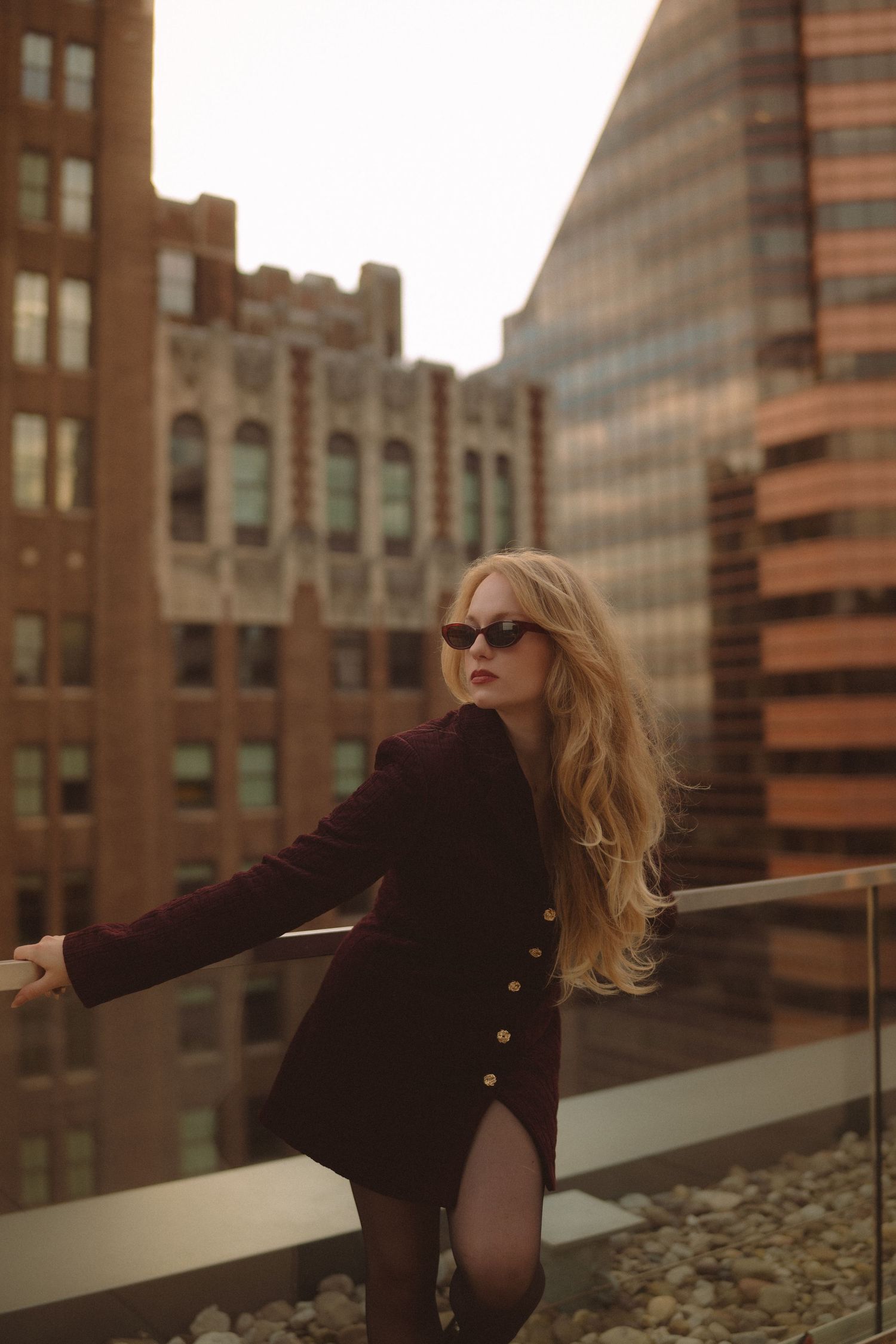 Blonde woman in dark blazer and sunglasses poses on urban rooftop balcony with city buildings backdrop.