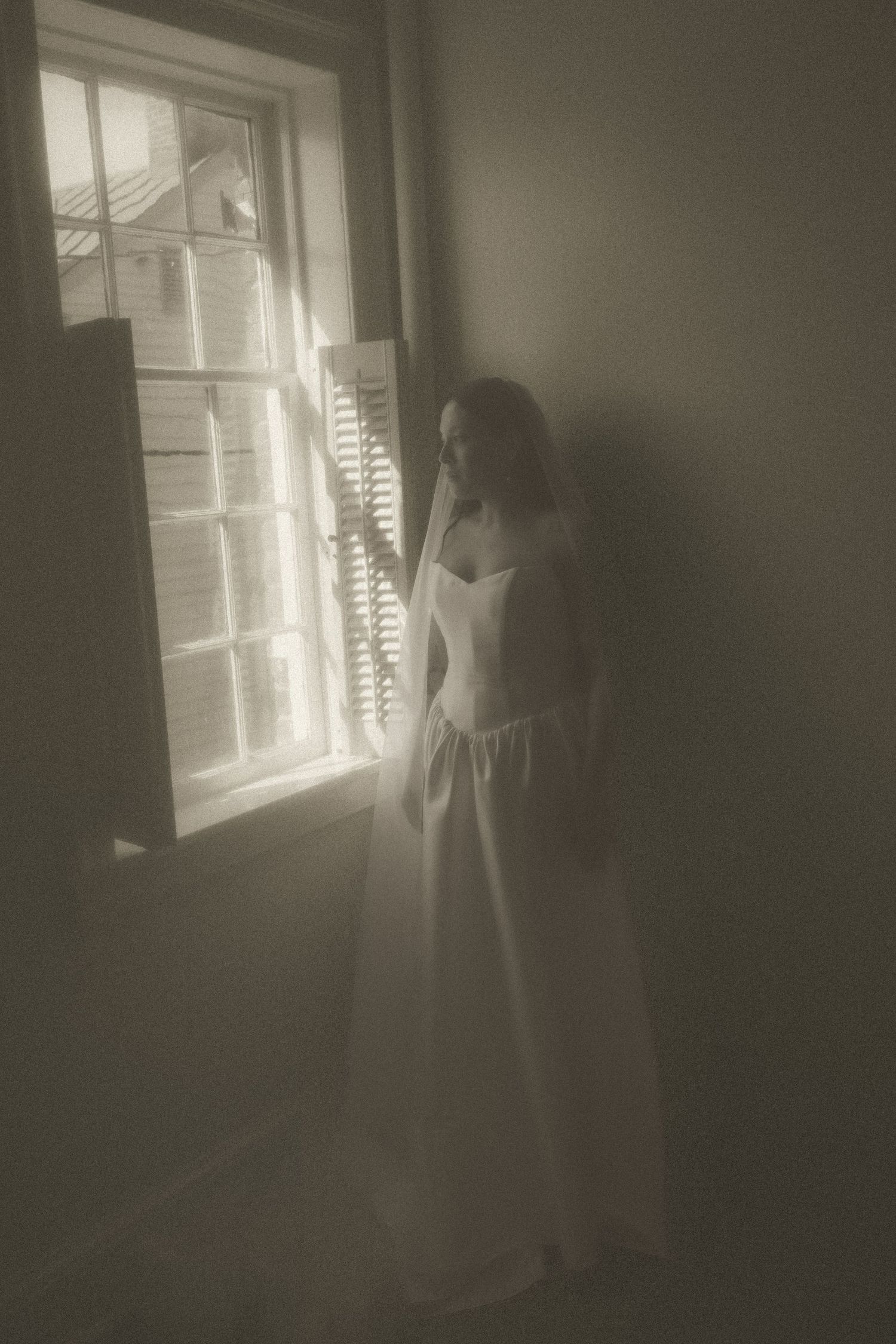 Woman in white dress standing by a sunlit window with shutters in a softly lit, dreamy monochrome interior.