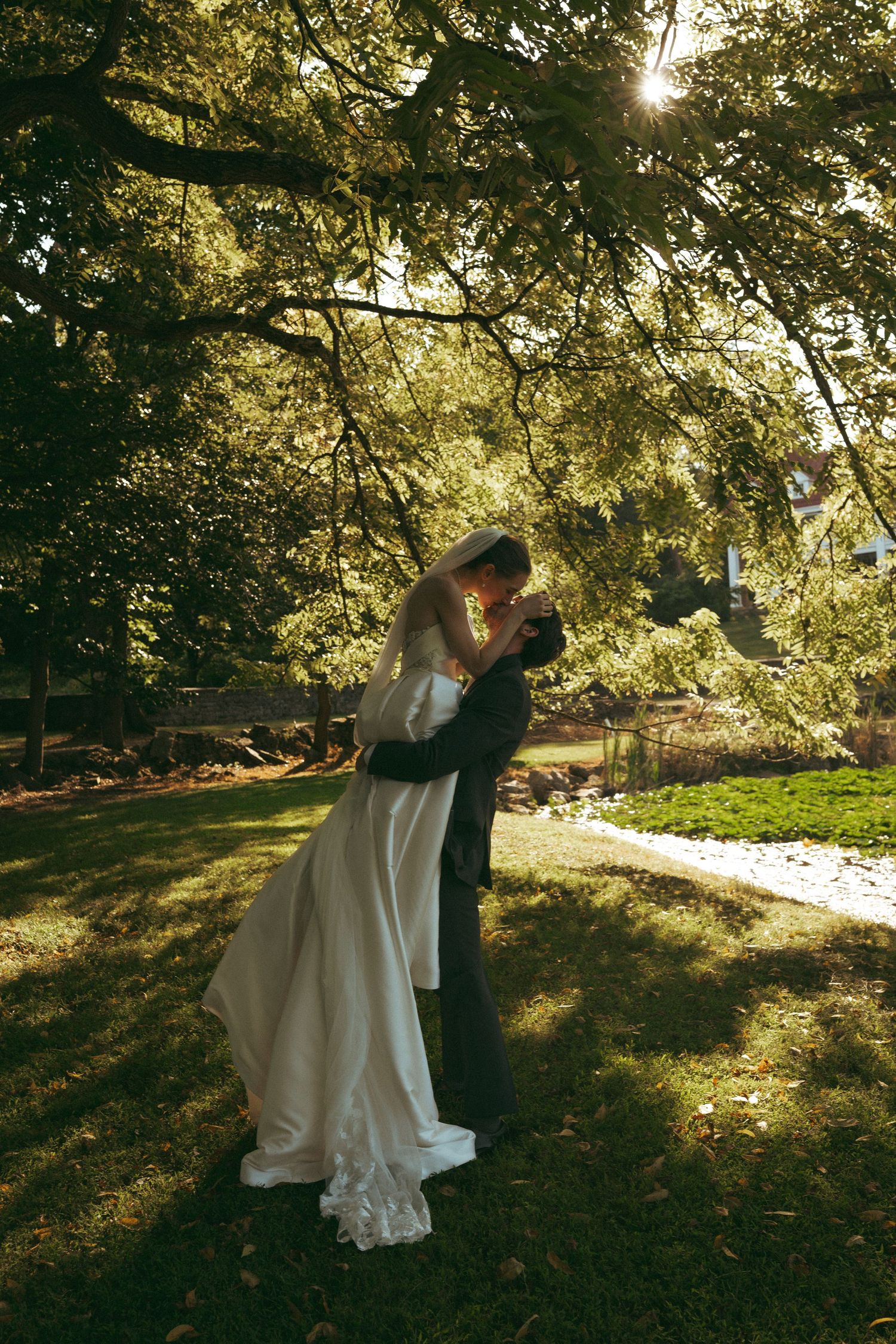 Bride and groom sharing a romantic kiss under lush green trees in a sunlit garden on their wedding day.