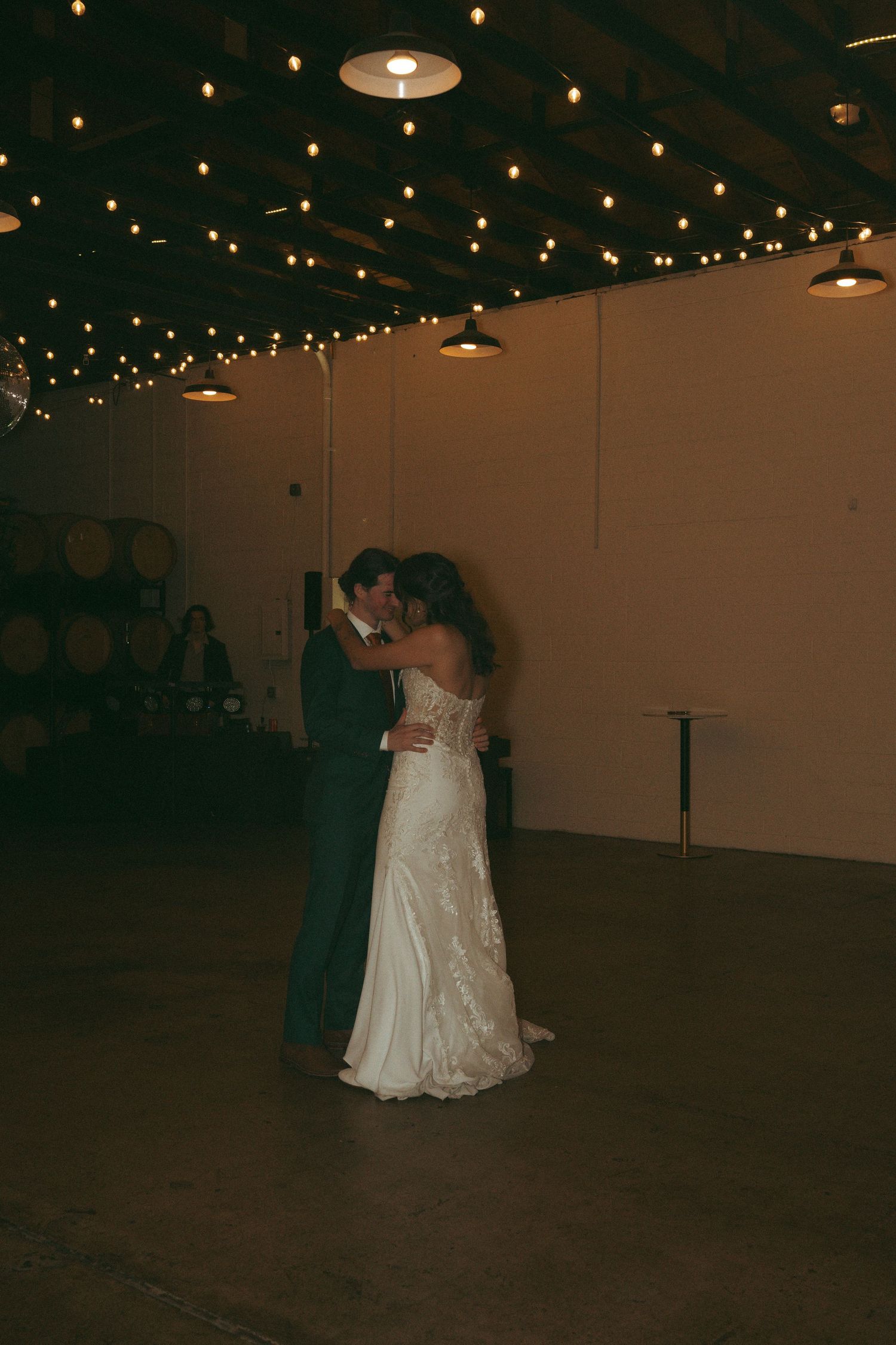 Bride and groom share first dance under warm string lights in a rustic winery wedding venue.