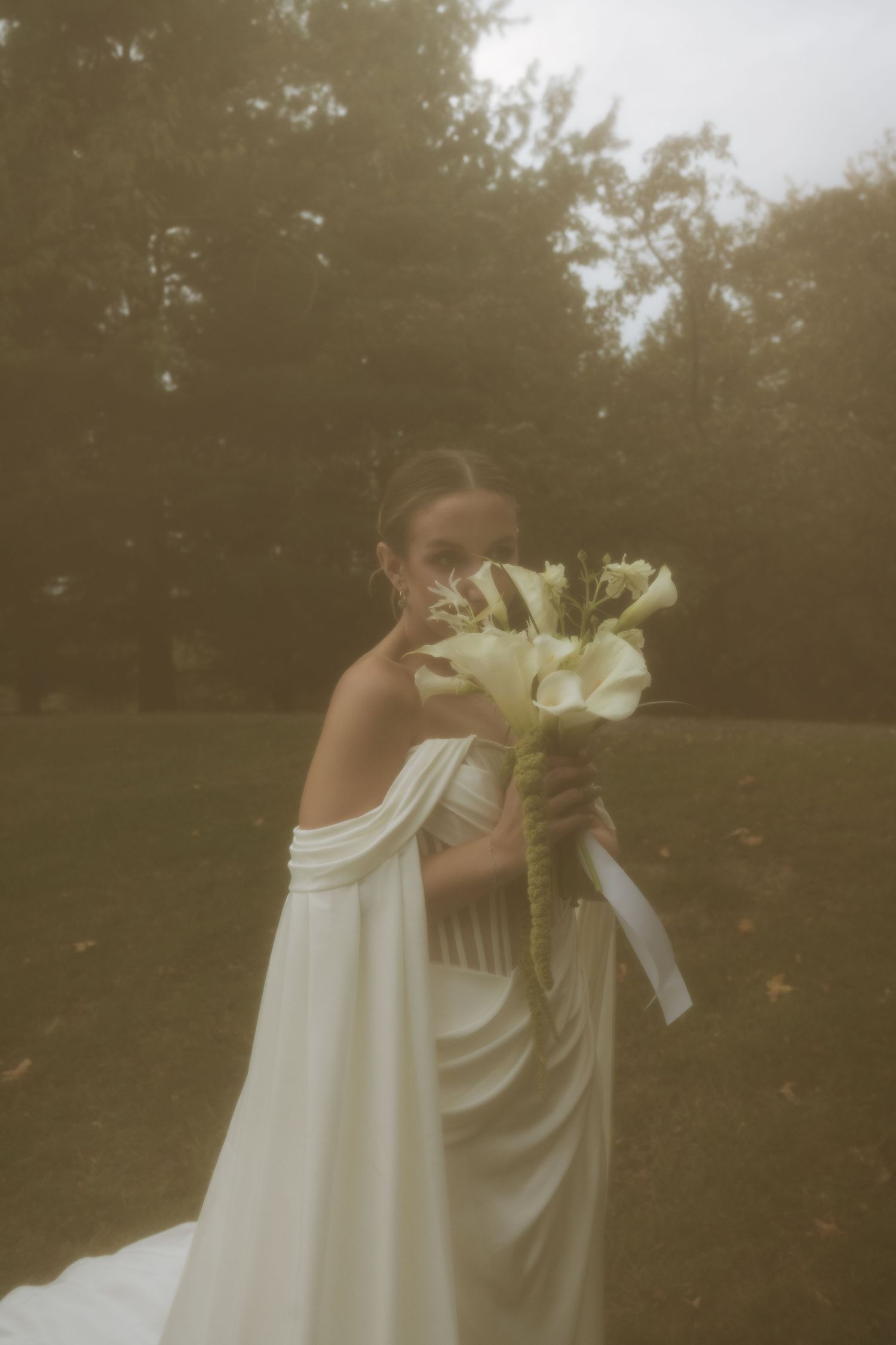 Bride in off-shoulder white gown holds elegant bouquet of white lilies outdoors in a lush green park setting.