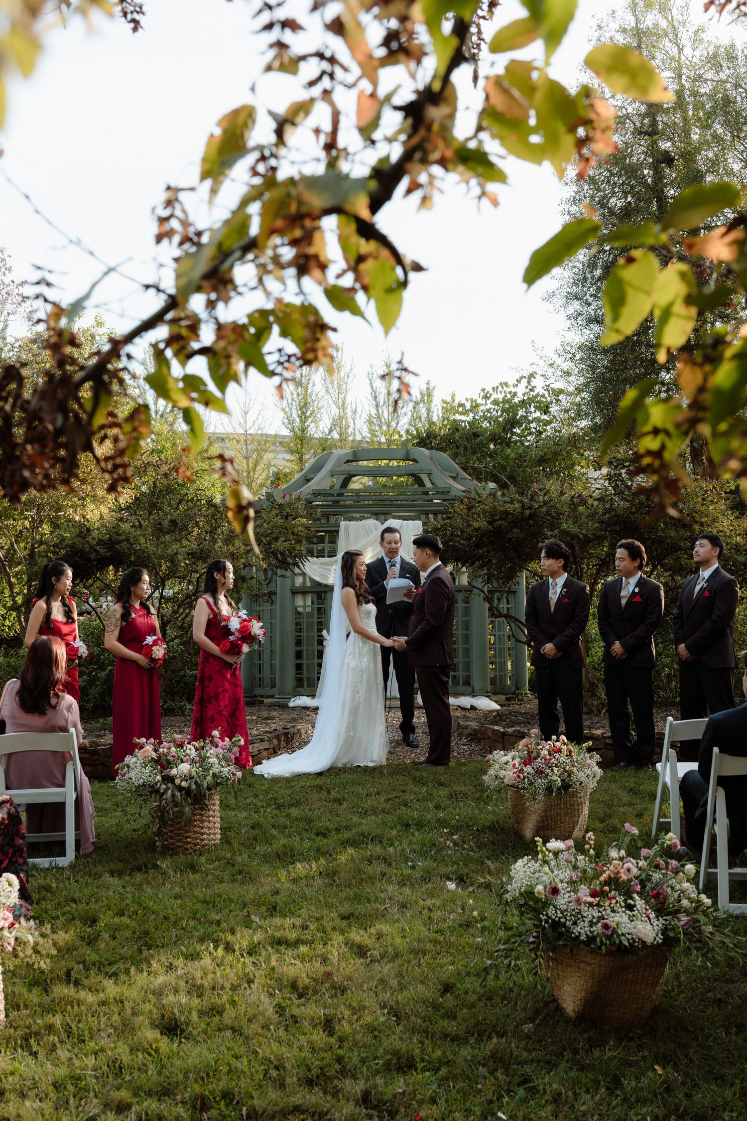 Outdoor garden wedding ceremony with bride and groom at altar, bridesmaids in red dresses, groomsmen, and floral arrangements.
