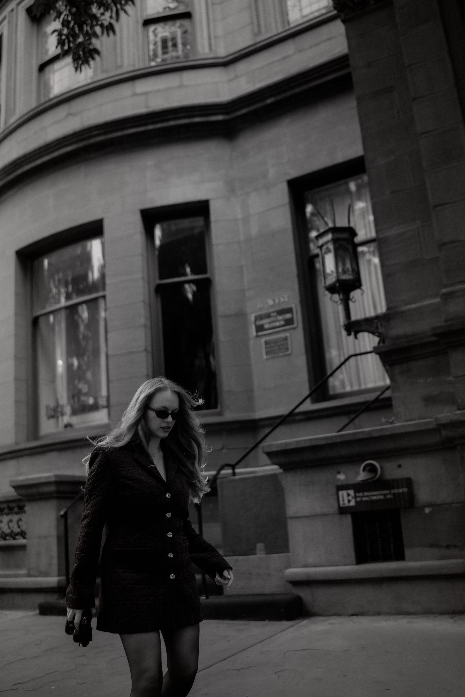Black and white photo of a blonde woman in a black mini dress walking on a city sidewalk past a stone building.
