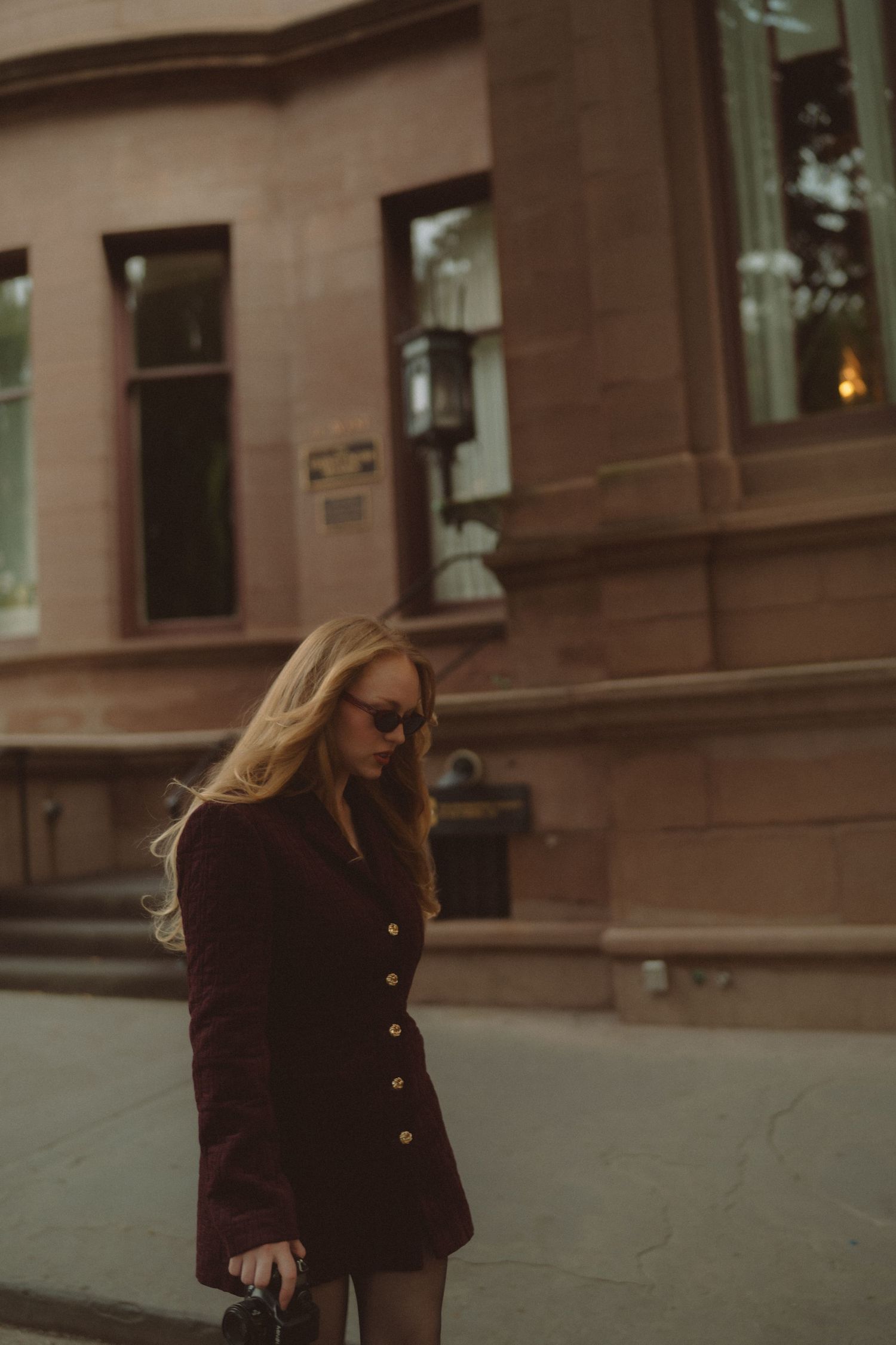 Blonde woman in dark burgundy blazer dress and sunglasses walking on a city sidewalk near a brownstone building.