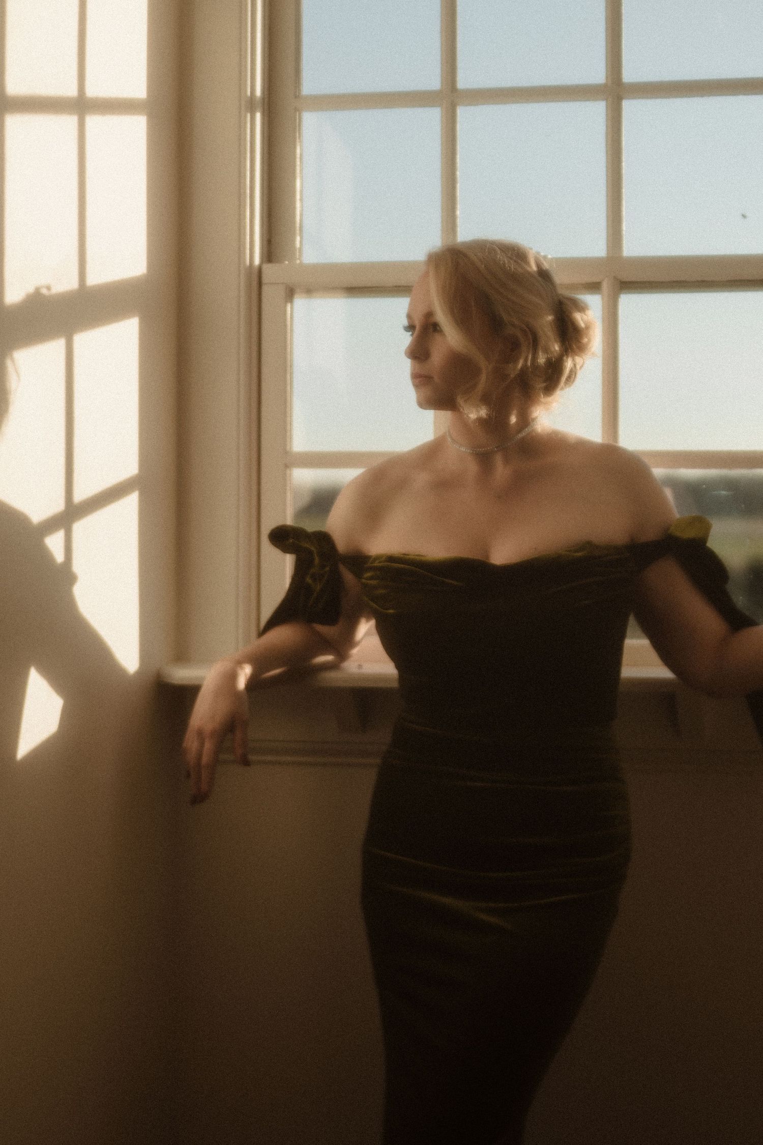 Elegant blonde woman in off-shoulder dark green dress gazing out sunlit window.