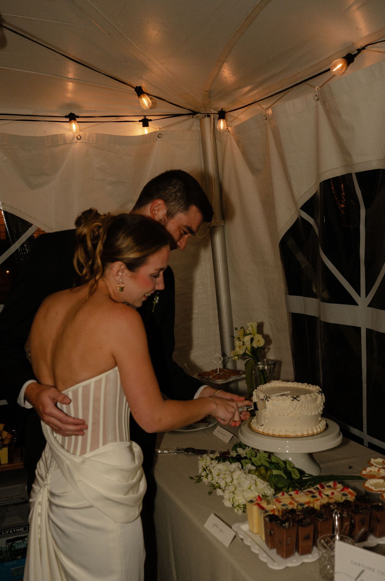 Bride and groom cutting their white wedding cake together under a tent with string lights at their reception.