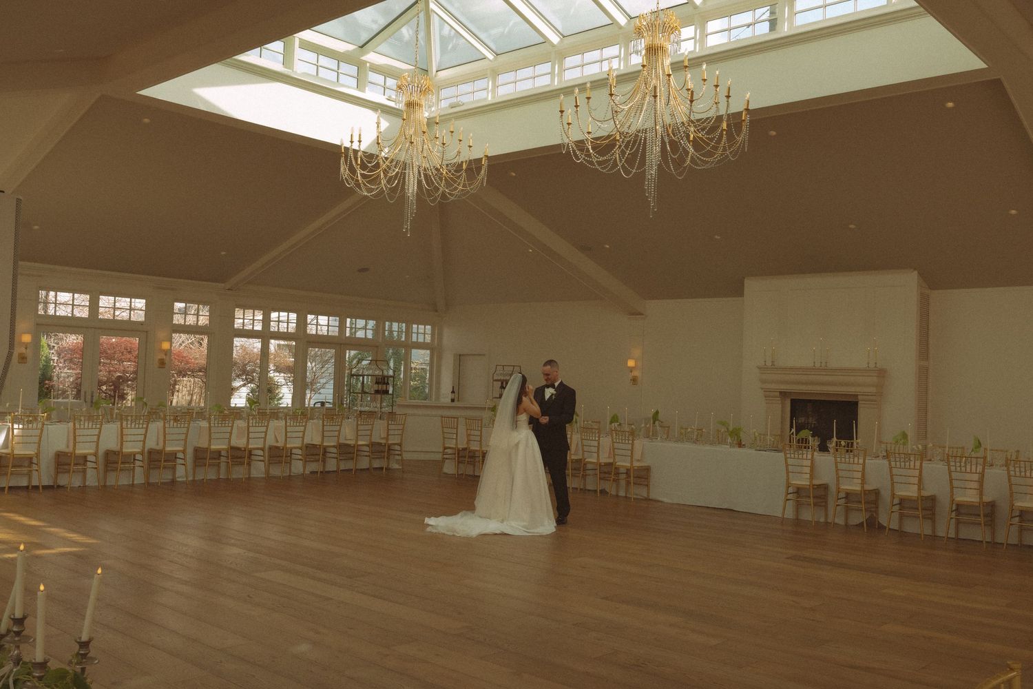 Bride and groom share first dance in elegant wedding venue with chandeliers, hardwood floors, and large windows.