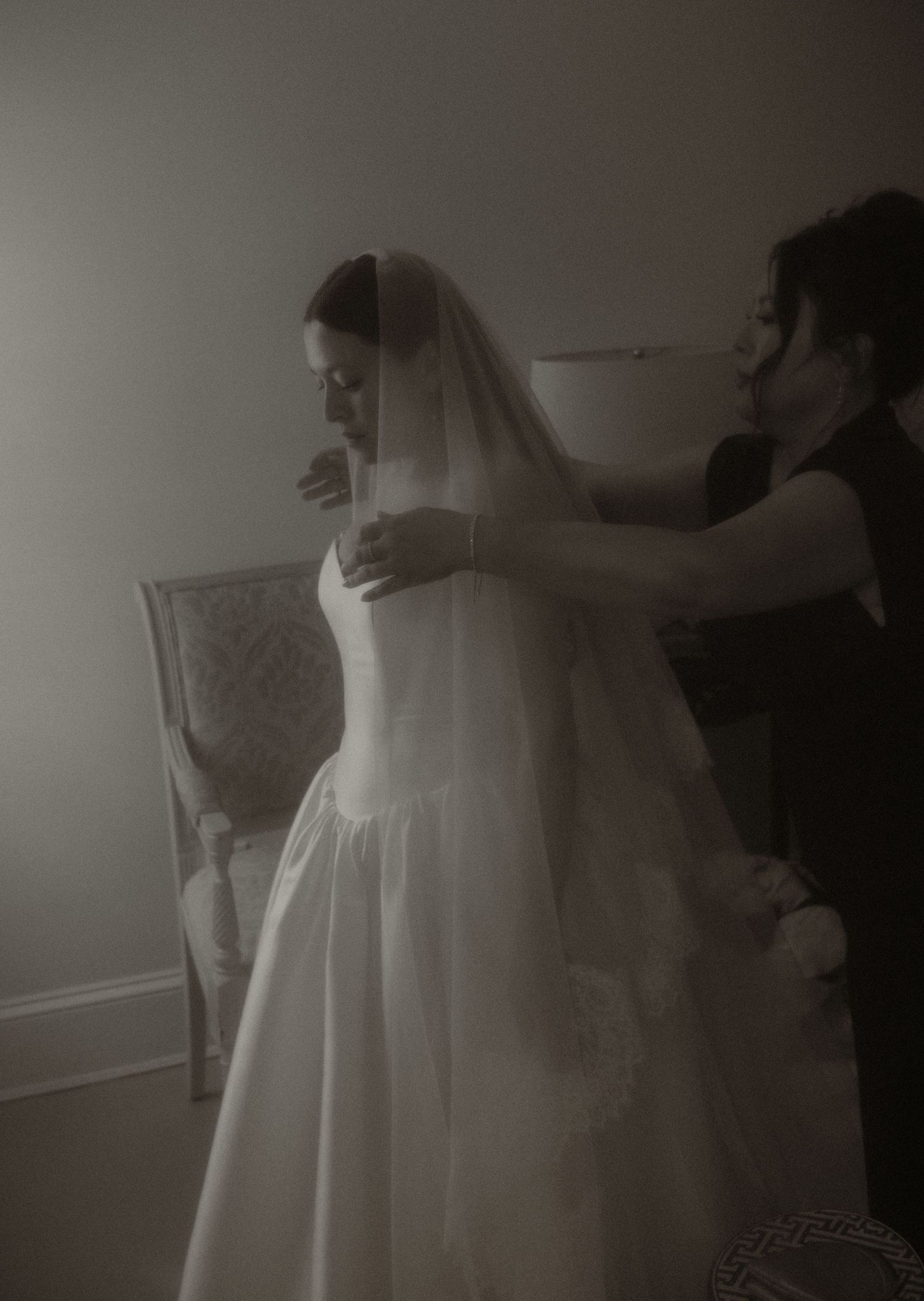 Black and white photo of a bride in a white gown and veil being helped by a bridesmaid before her wedding.