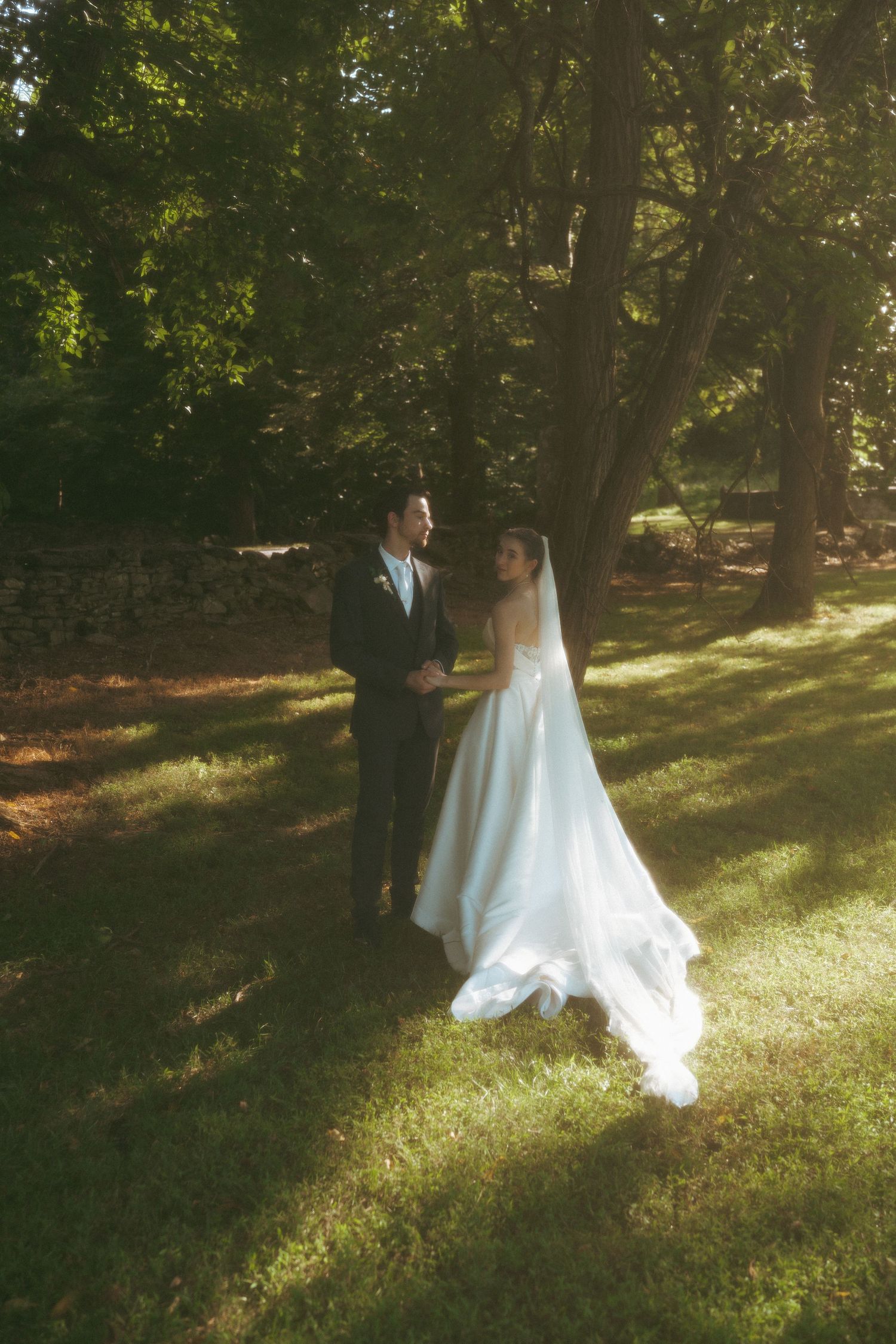 Bride in white gown and groom in dark suit hold hands in sunlit garden surrounded by lush green trees.
