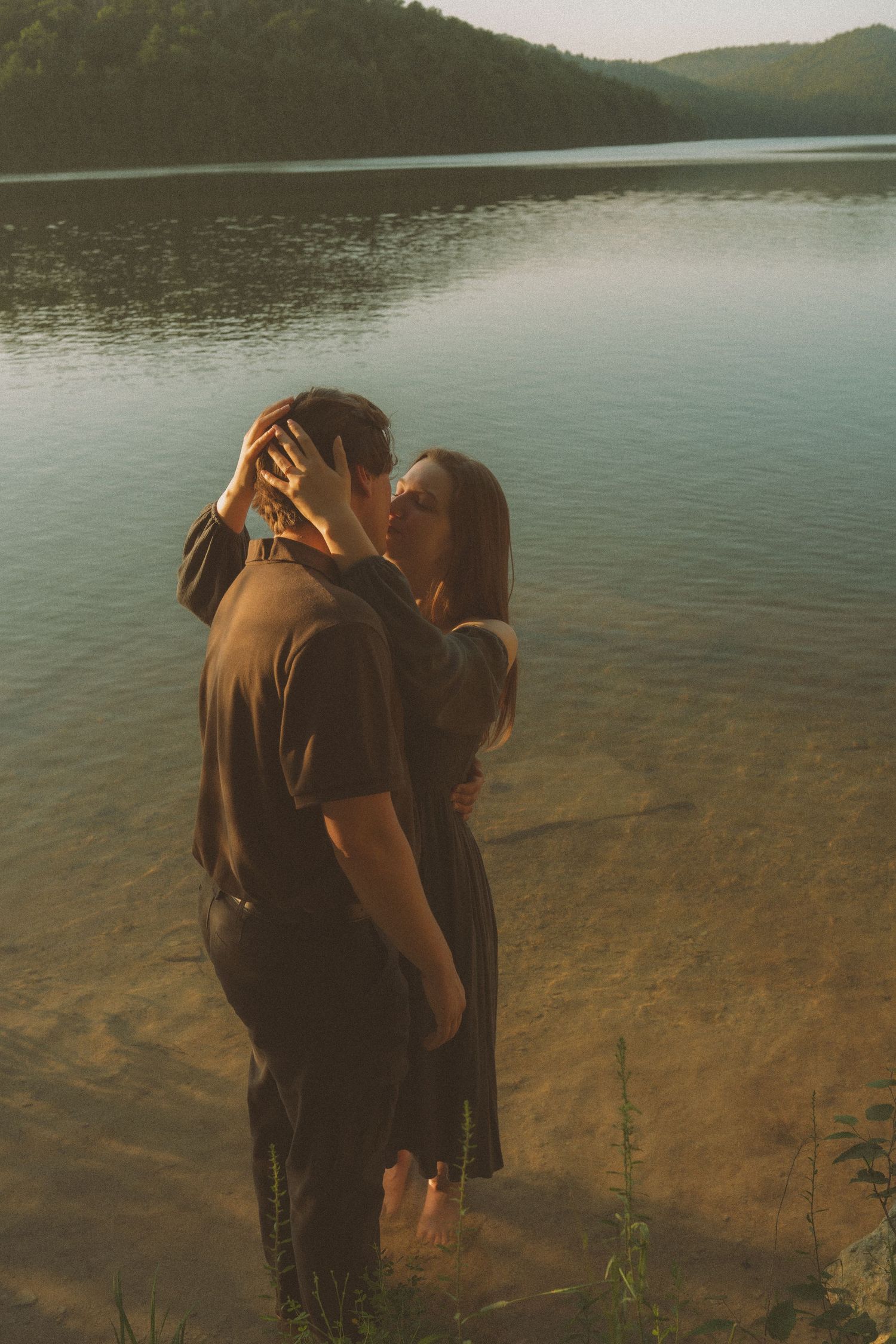 Couple sharing a romantic kiss while standing in shallow lake water at golden hour with lush green hills in background.