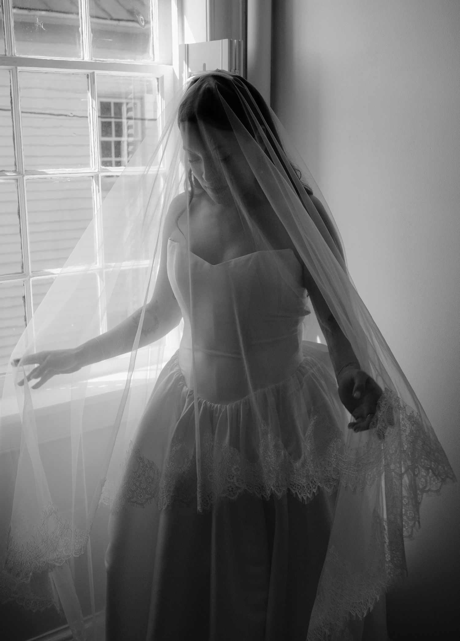 Black and white photo of a bride in a strapless wedding dress and lace veil standing by a window.