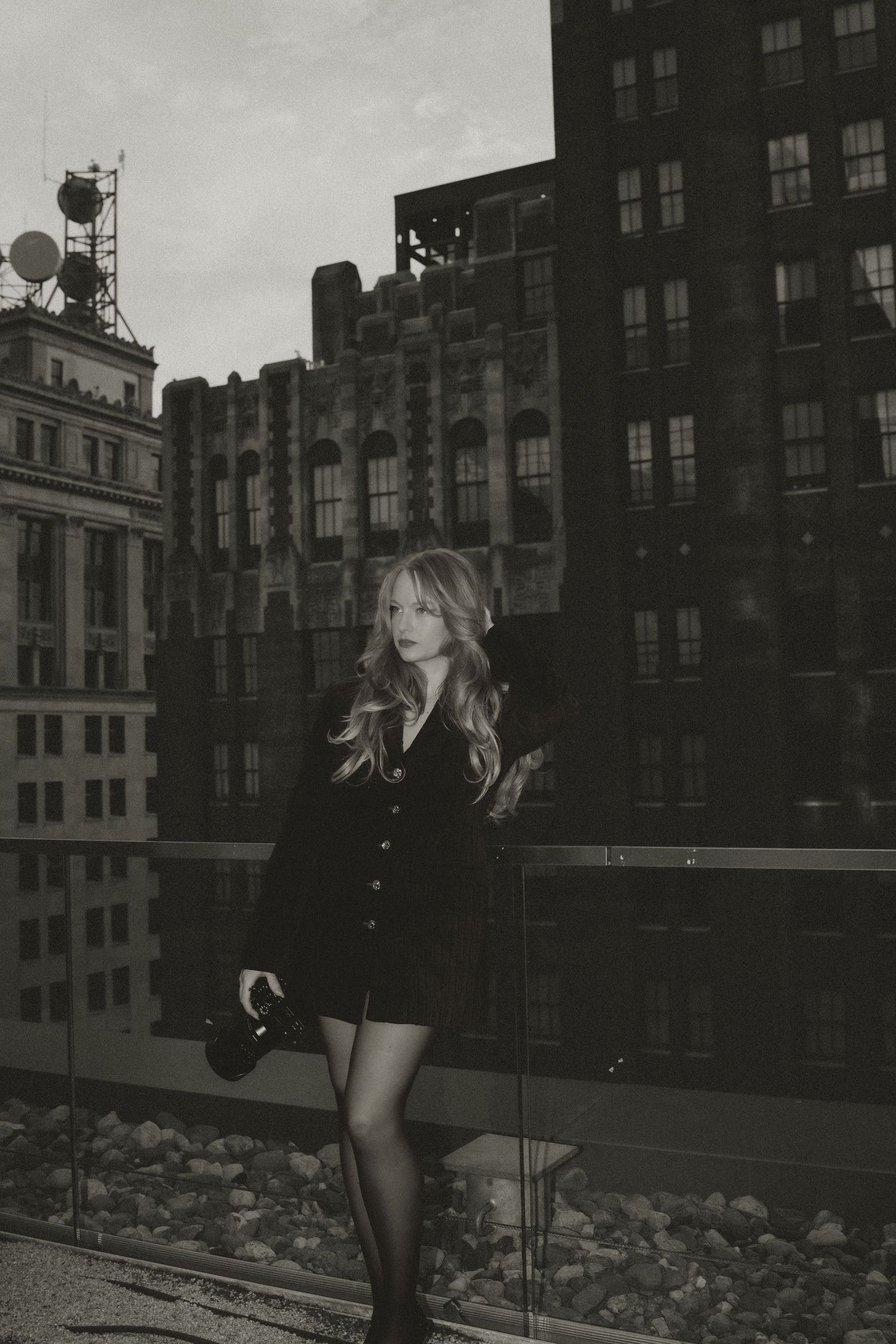 Black and white photo of a blonde woman in a dark dress posing on a rooftop with city buildings in the background.