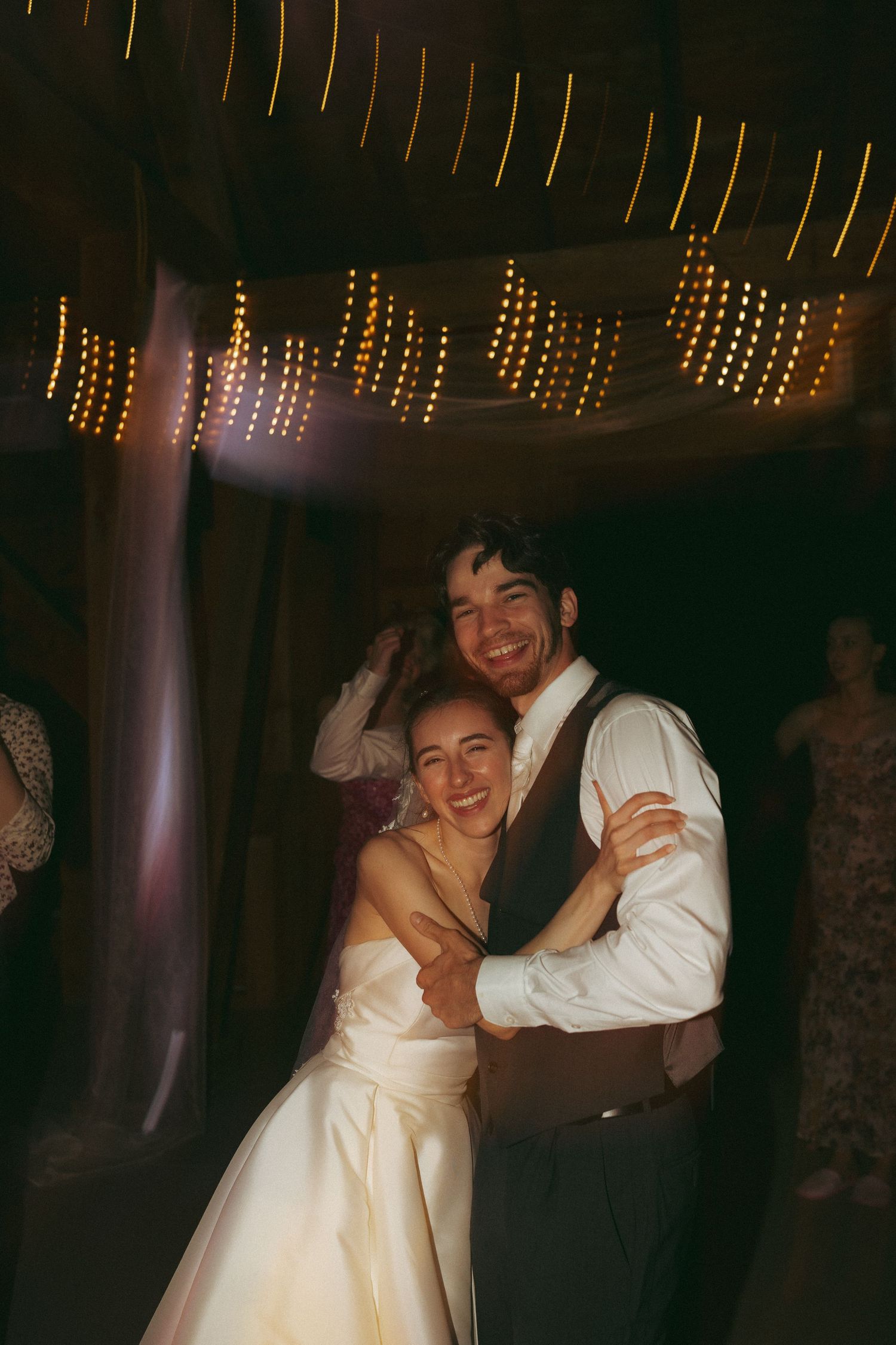 Bride and groom share a joyful embrace on the dance floor under warm string lights at their wedding reception.