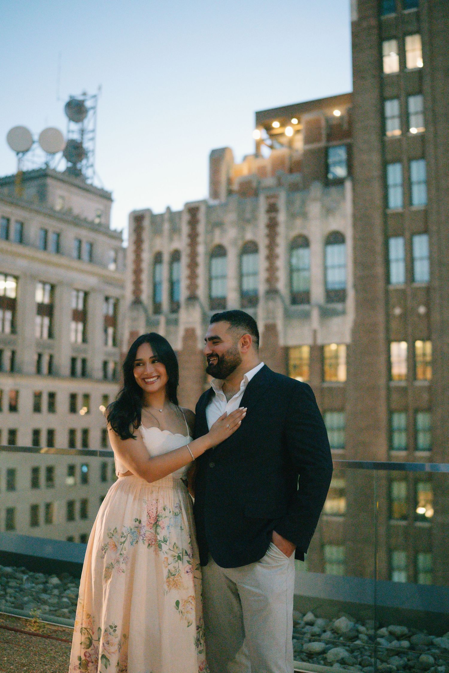 Couple poses on urban rooftop at dusk, woman in floral gown smiling, man in navy blazer, city buildings backdrop.