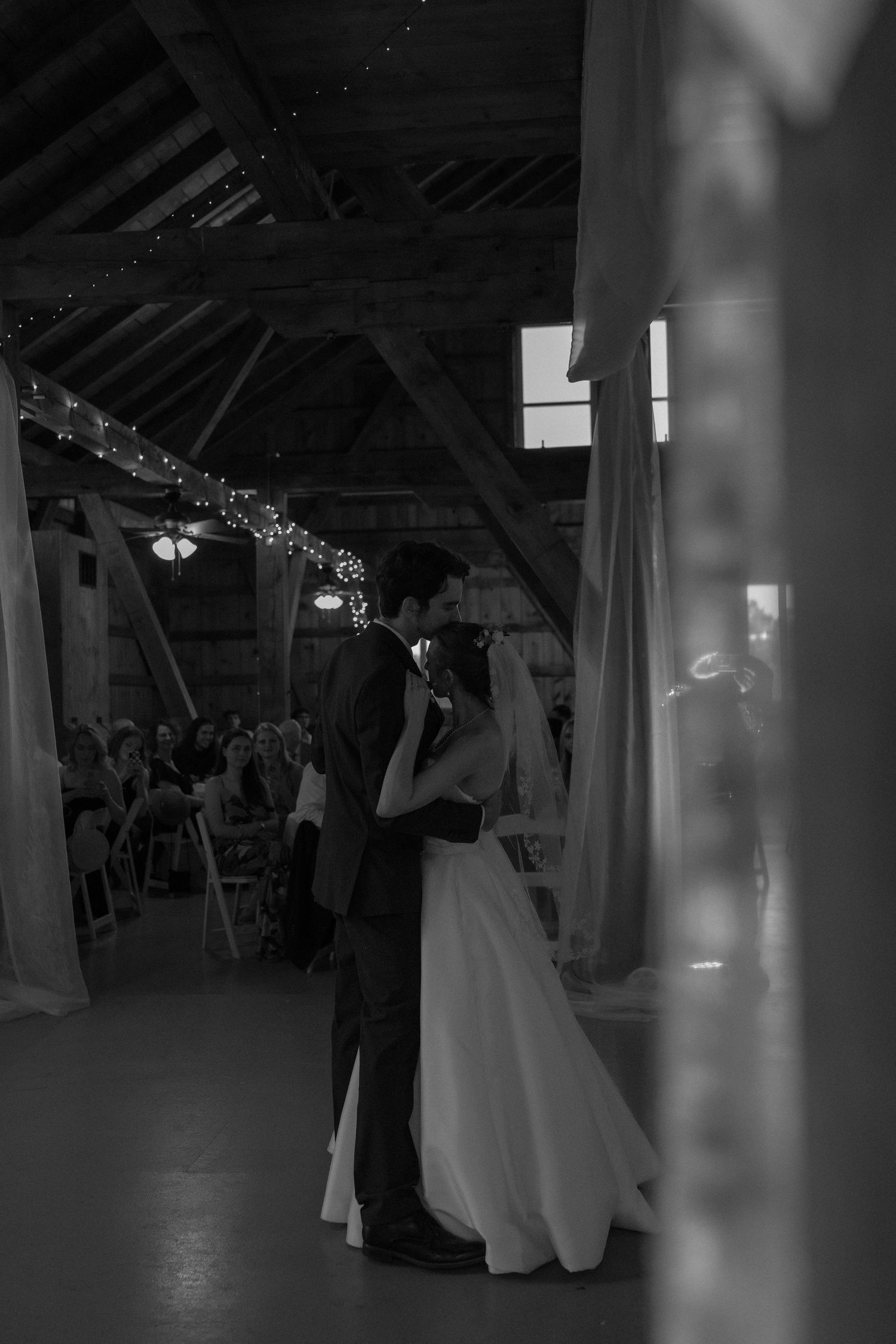 Black and white photo of bride and groom sharing first dance in rustic barn venue with string lights.
