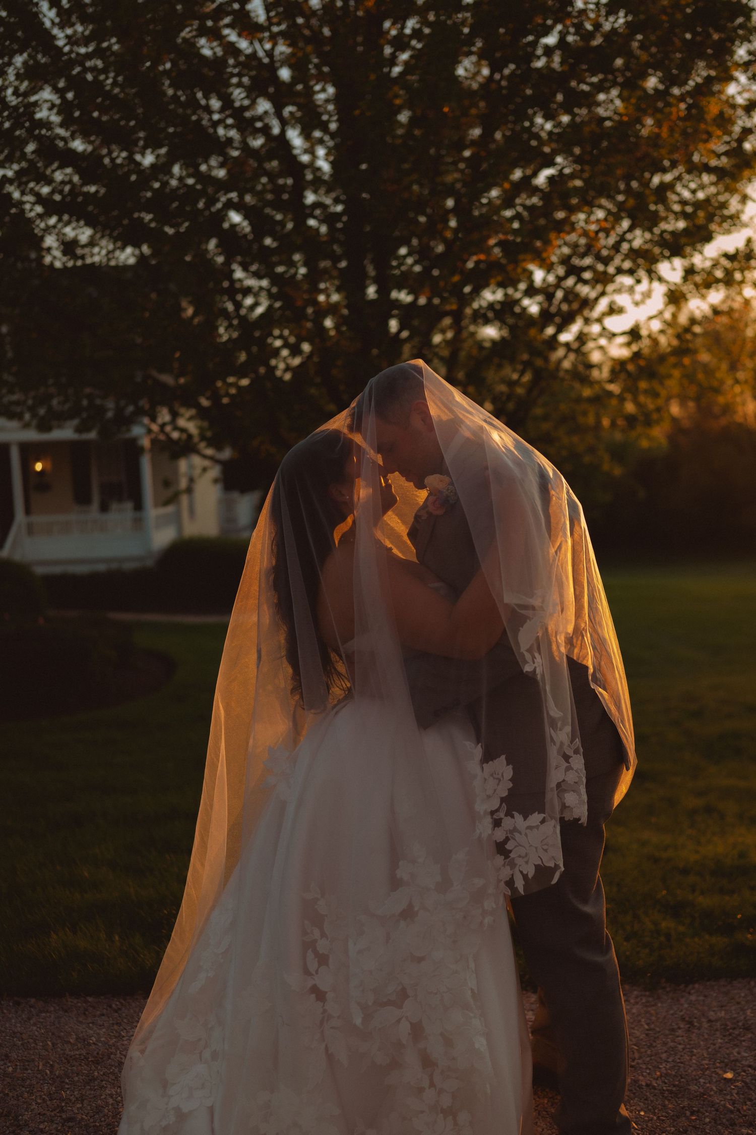 Bride and groom sharing a kiss under a flowing veil at golden hour sunset outdoors.