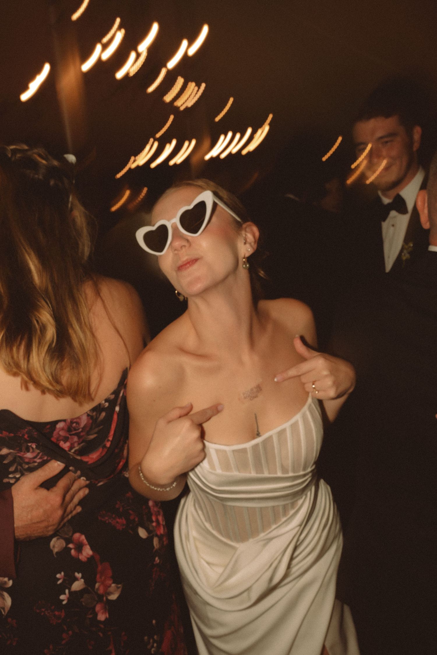 Bride dancing in white gown and heart-shaped sunglasses at wedding reception with warm bokeh lights.