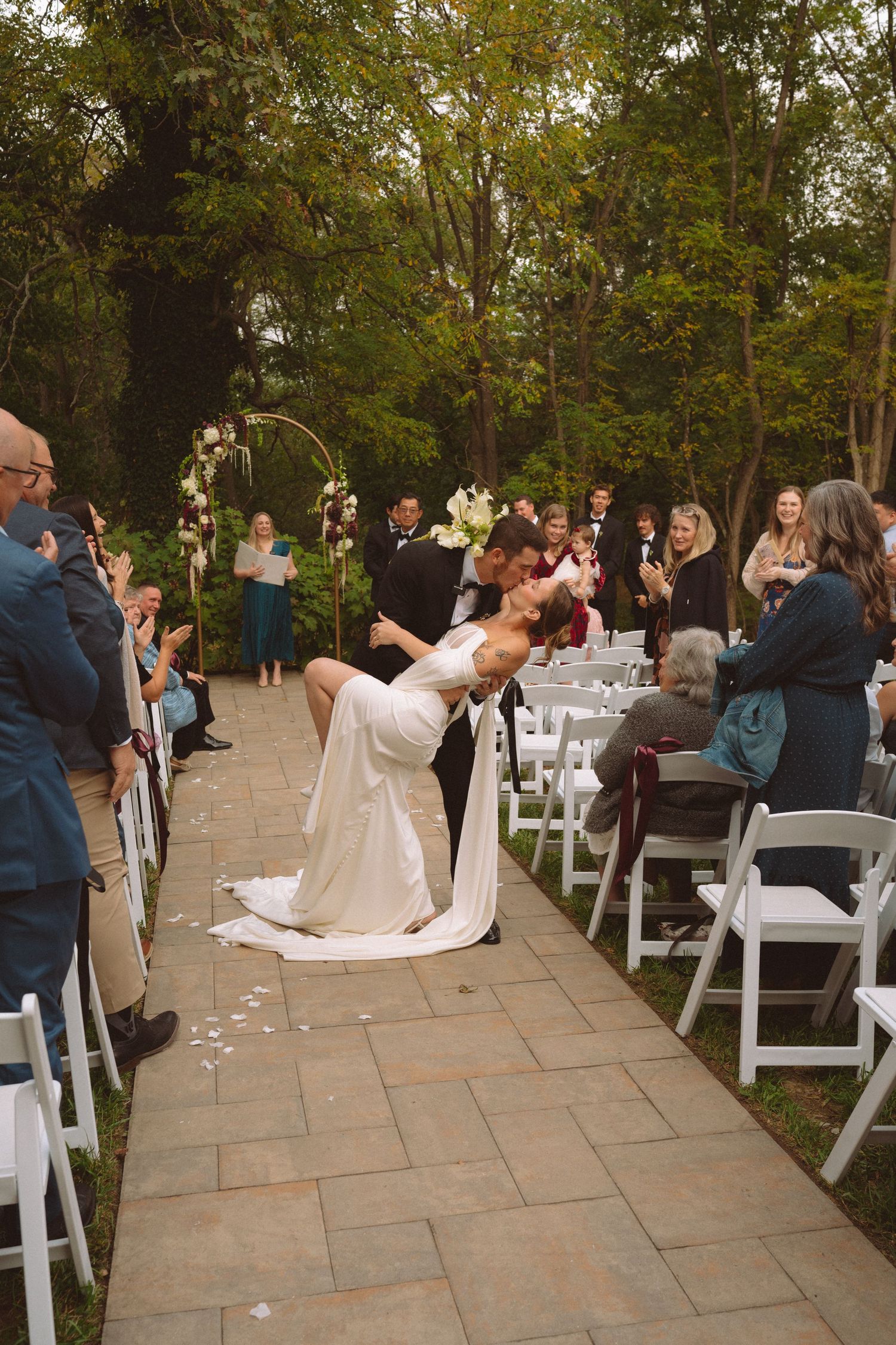 Groom dips bride in a romantic kiss while walking down the aisle at an outdoor wedding ceremony.