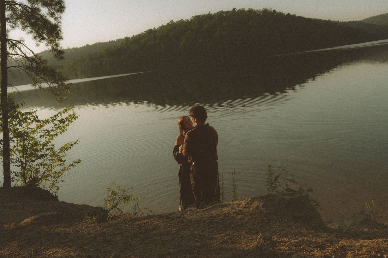 Couple embracing at golden hour by a serene lake with forested hills in the background.