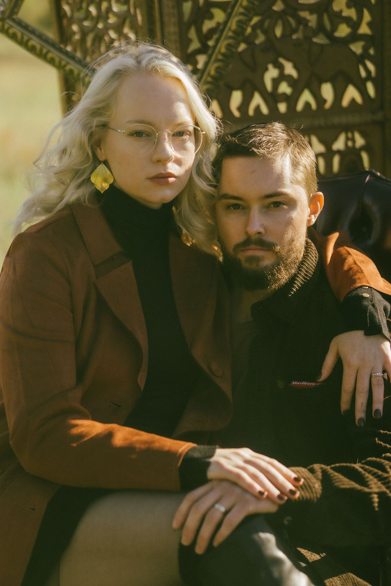 A blonde woman in glasses and brown coat sits with a bearded man outdoors on an ornate metal bench.
