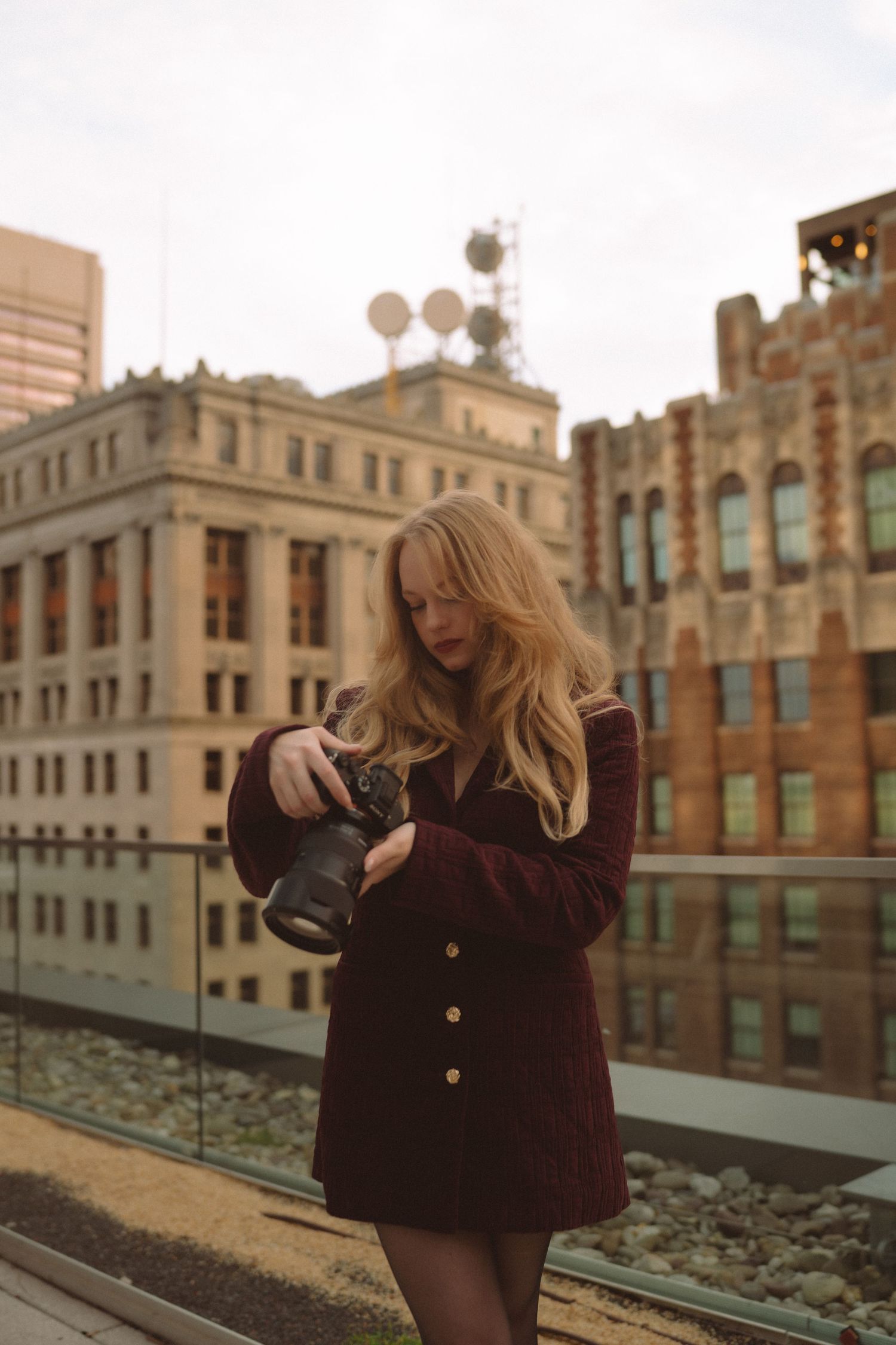 A blonde woman in a burgundy coat reviews photos on her camera on a rooftop with urban buildings behind her.