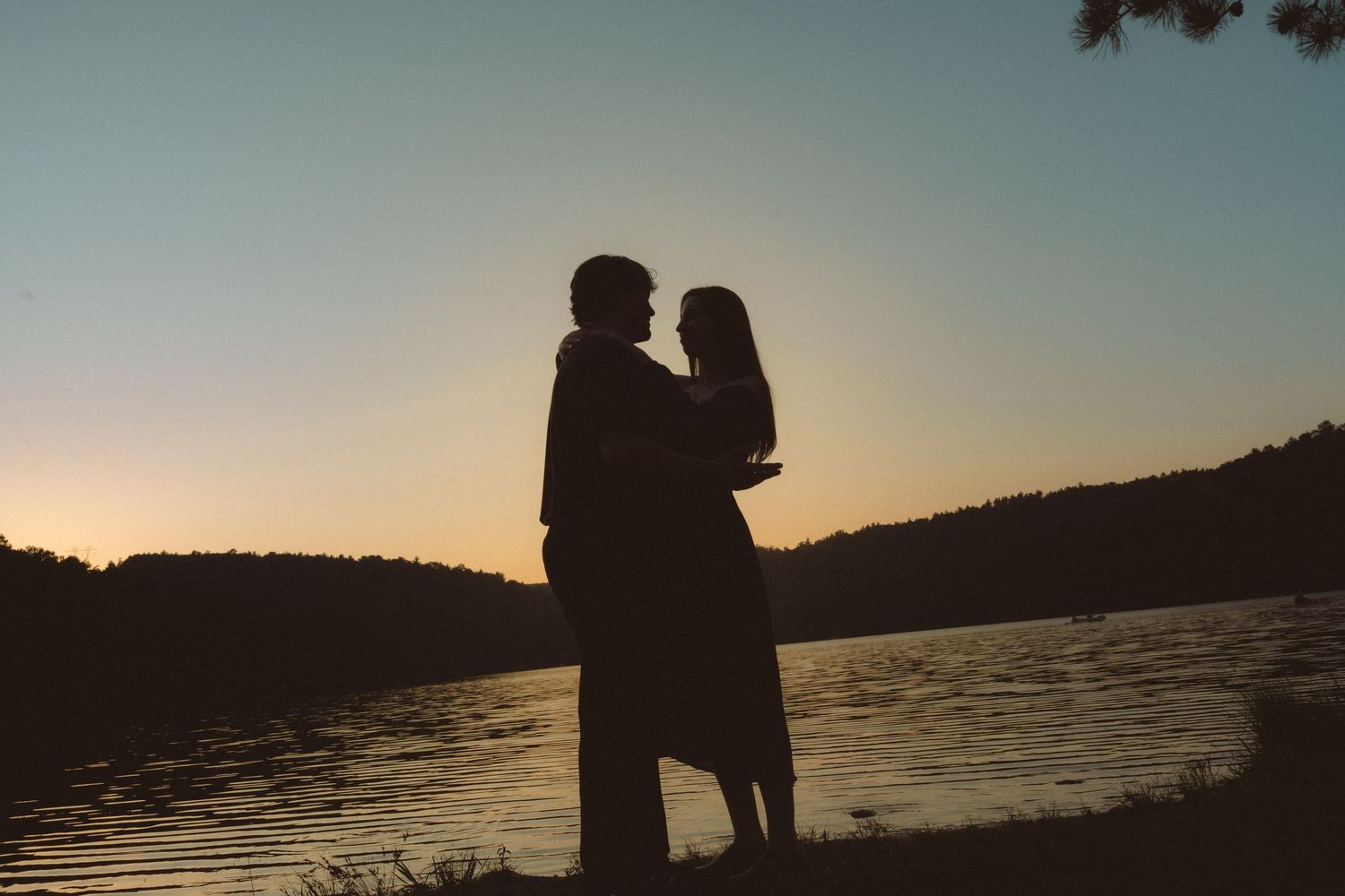 Silhouette of a romantic couple embracing by a lake at sunset with golden sky and tree-lined hills.