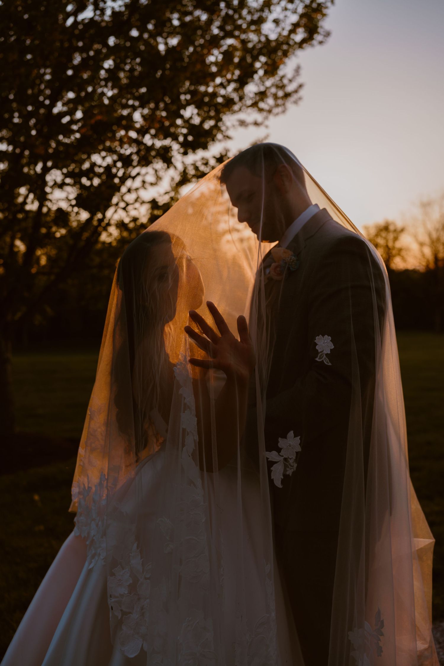 Bride and groom share romantic sunset moment under wedding veil, golden hour backlight, outdoor ceremony.