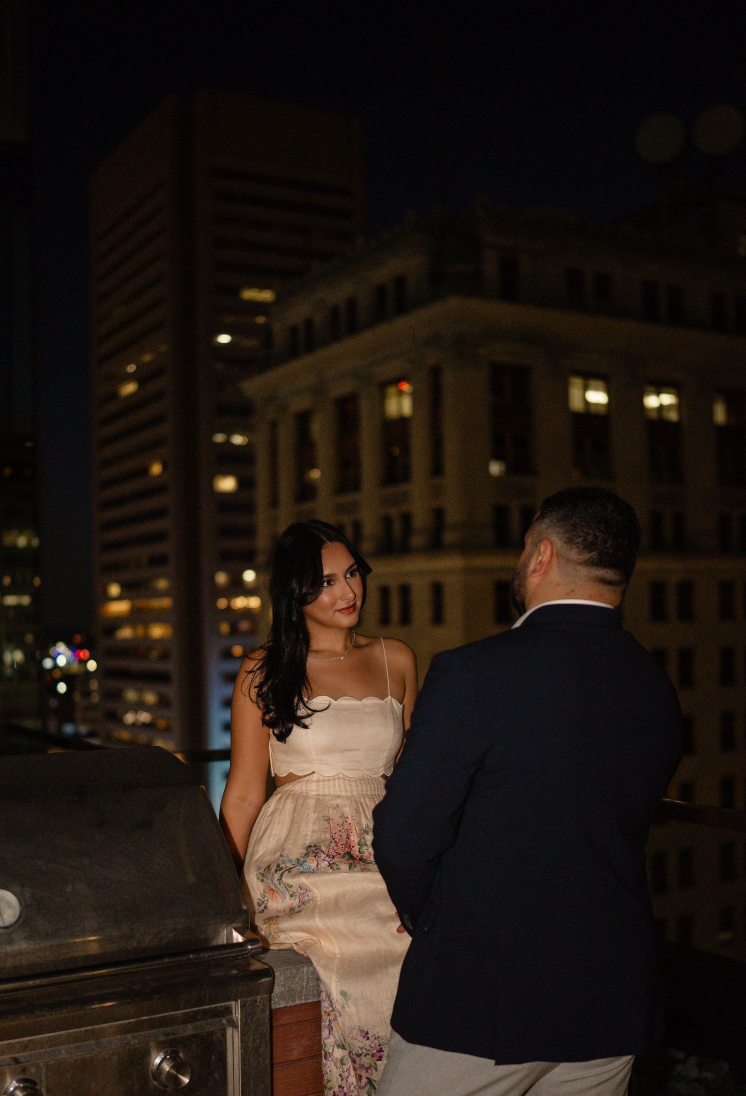 Couple sharing a romantic moment on a rooftop at night with city buildings glowing in the background.