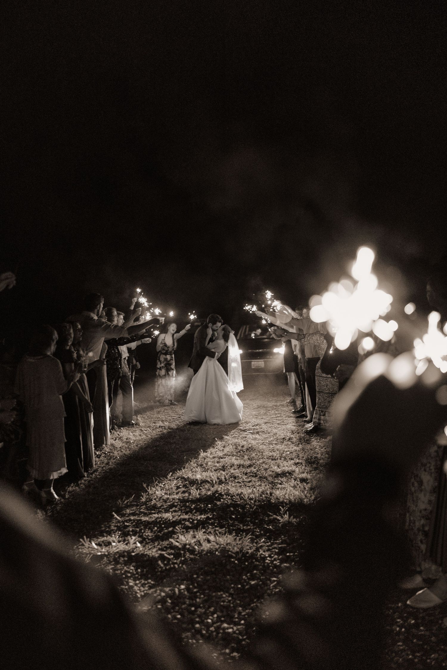 Bride and groom walk through a sparkler exit tunnel at night in a romantic black and white wedding photo.