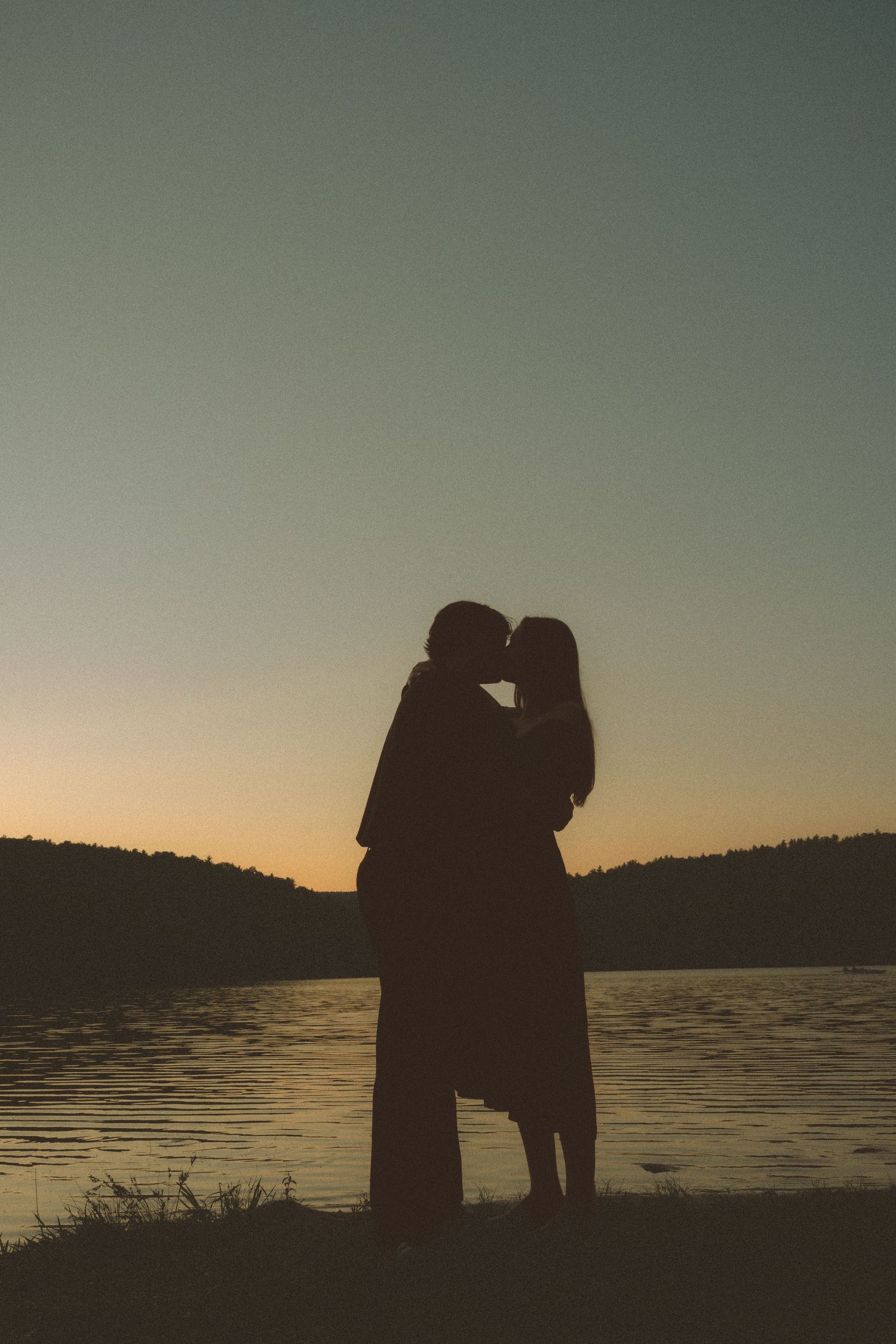 Silhouette of a couple kissing by a lake at sunset with warm golden sky and dark treeline.