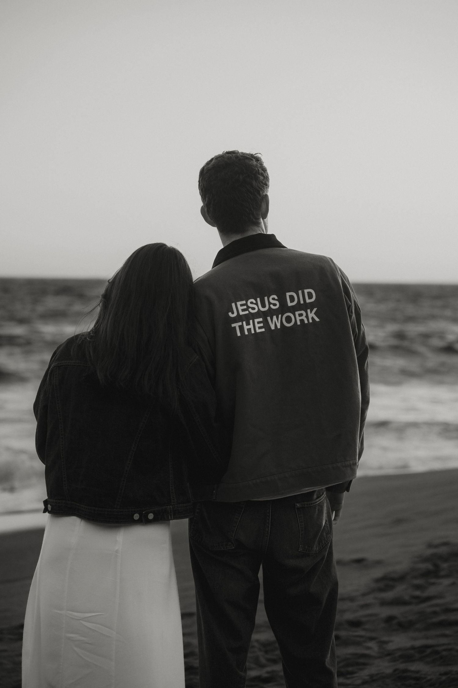 Couple standing together on beach, man wearing Jesus Did The Work jacket, black and white photo.